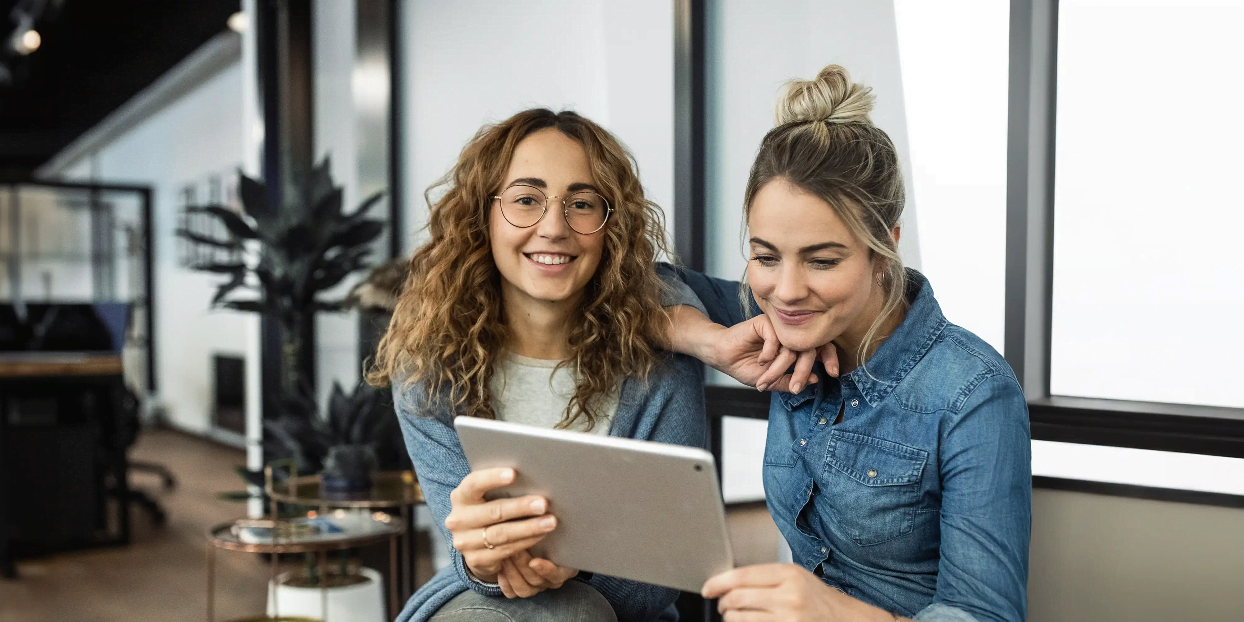 Zwei Frauen sitzen in einem Büro und schauen gemeinsam lächelnd auf ein Tablet. Beide wirken entspannt und konzentriert. Die moderne Büroeinrichtung im Hintergrund vermittelt eine produktive und freundliche Arbeitsatmosphäre.
