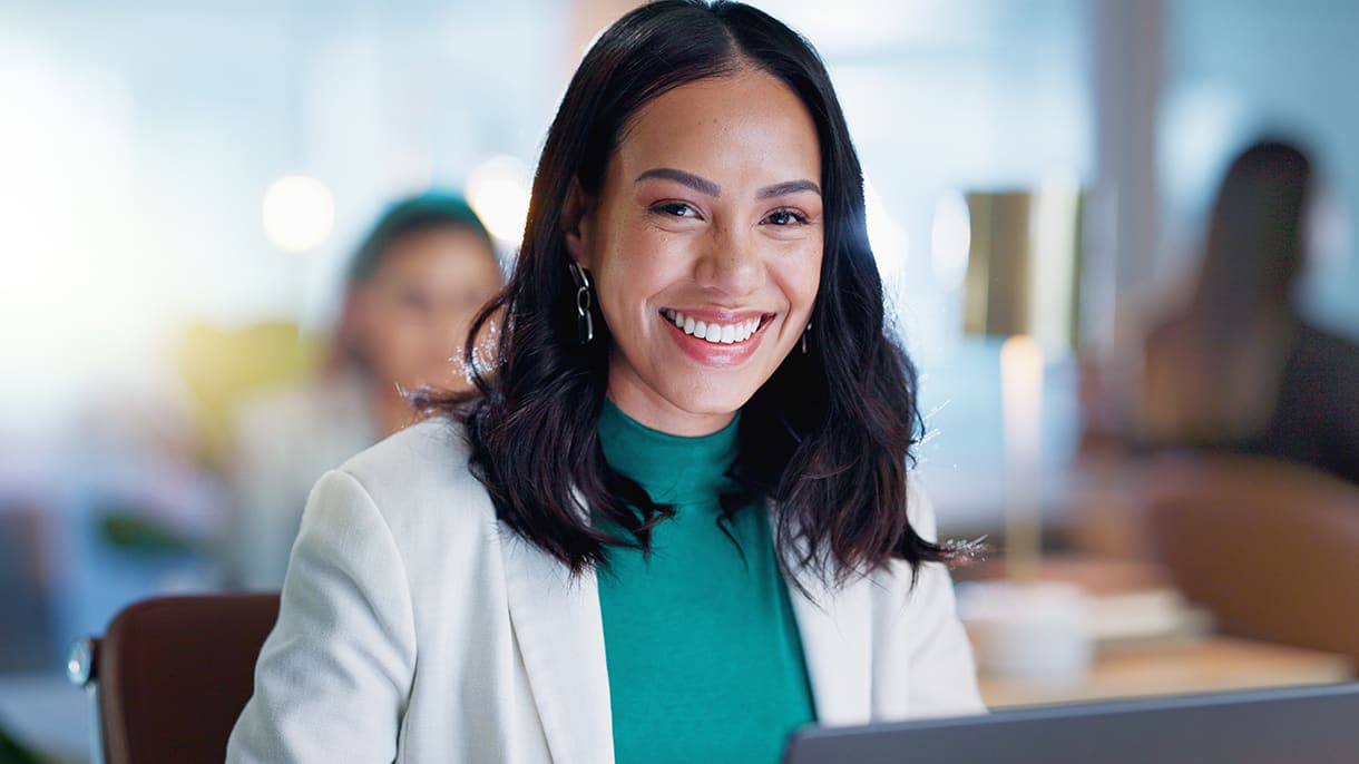 A smiling woman in a professional setting uses a laptop, representing the flexibility and engagement of online MBA courses.