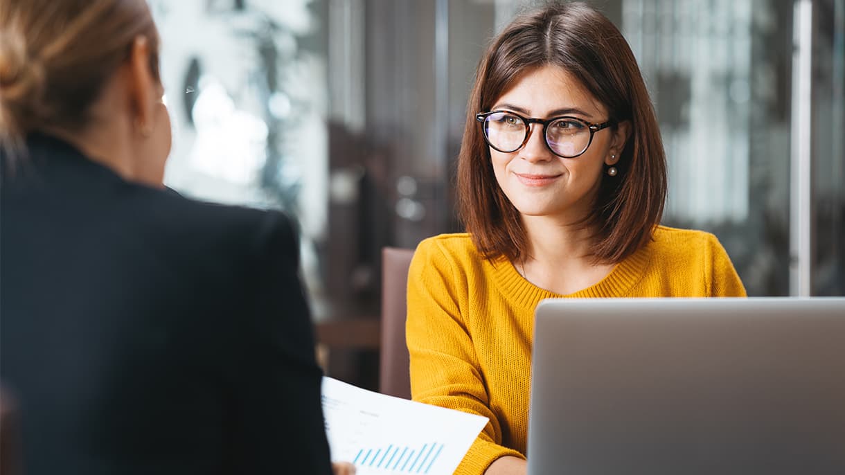 A smiling woman in glasses sits at a desk with a laptop, engaged in a discussion with another person, representing opportunities and support for international students.