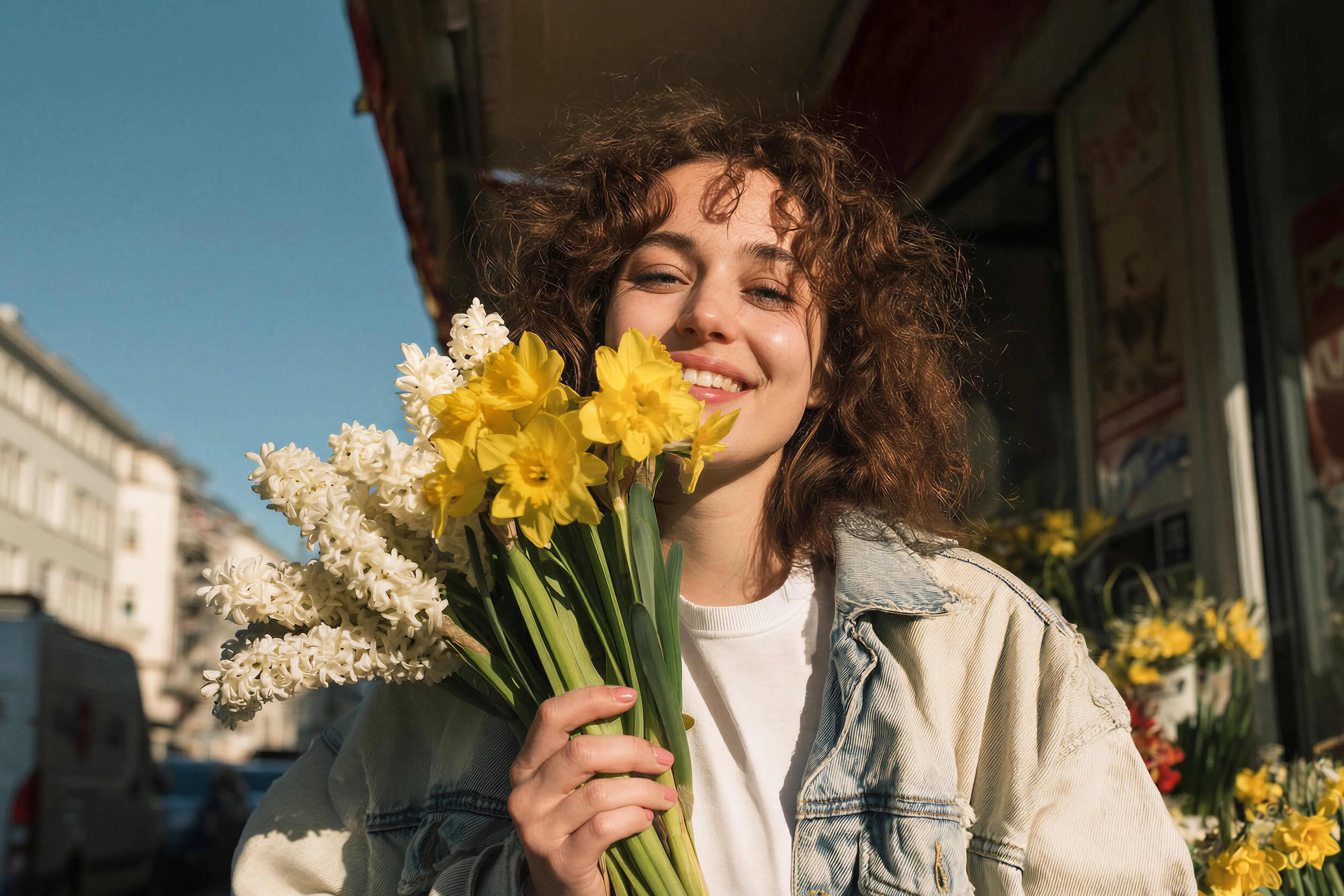 Lächelnde Frau mit lockigem Haar hält einen Strauß gelber und weißer Frühlingsblumen vor städtischem Hintergrund
