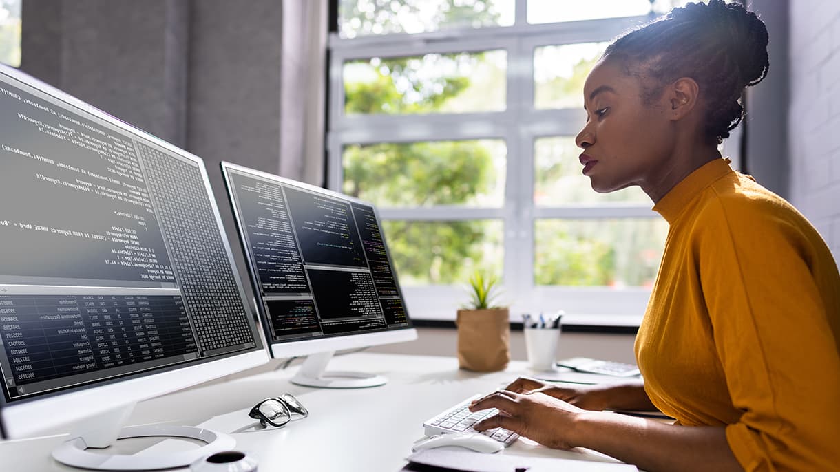 A person in a mustard yellow shirt working on multiple computer screens with code in a bright office space, illustrating living abroad in Berlin while pursuing a tech career.