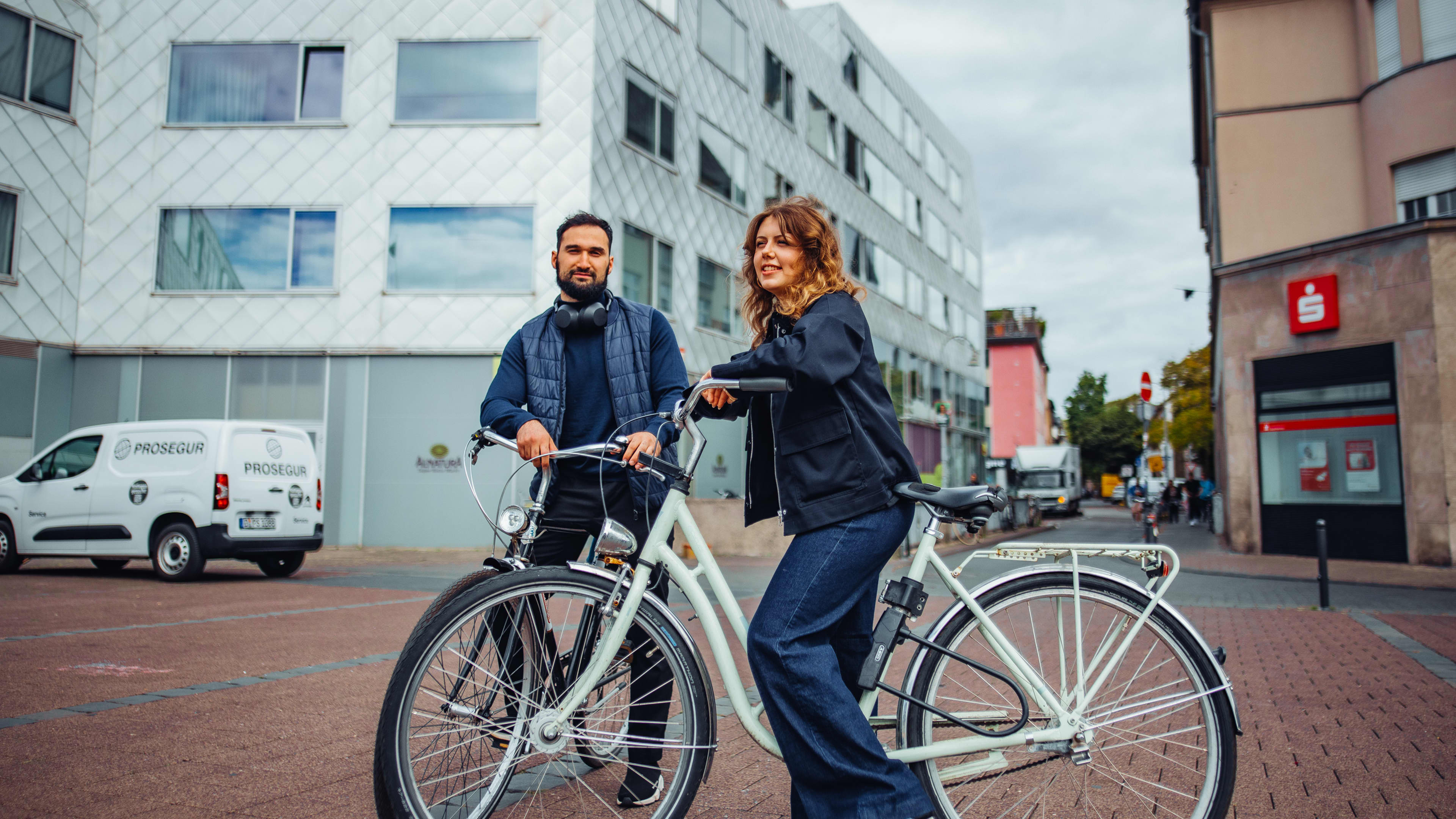 Two students standing with their bicycles on a Cologne city street in front of a modern building.