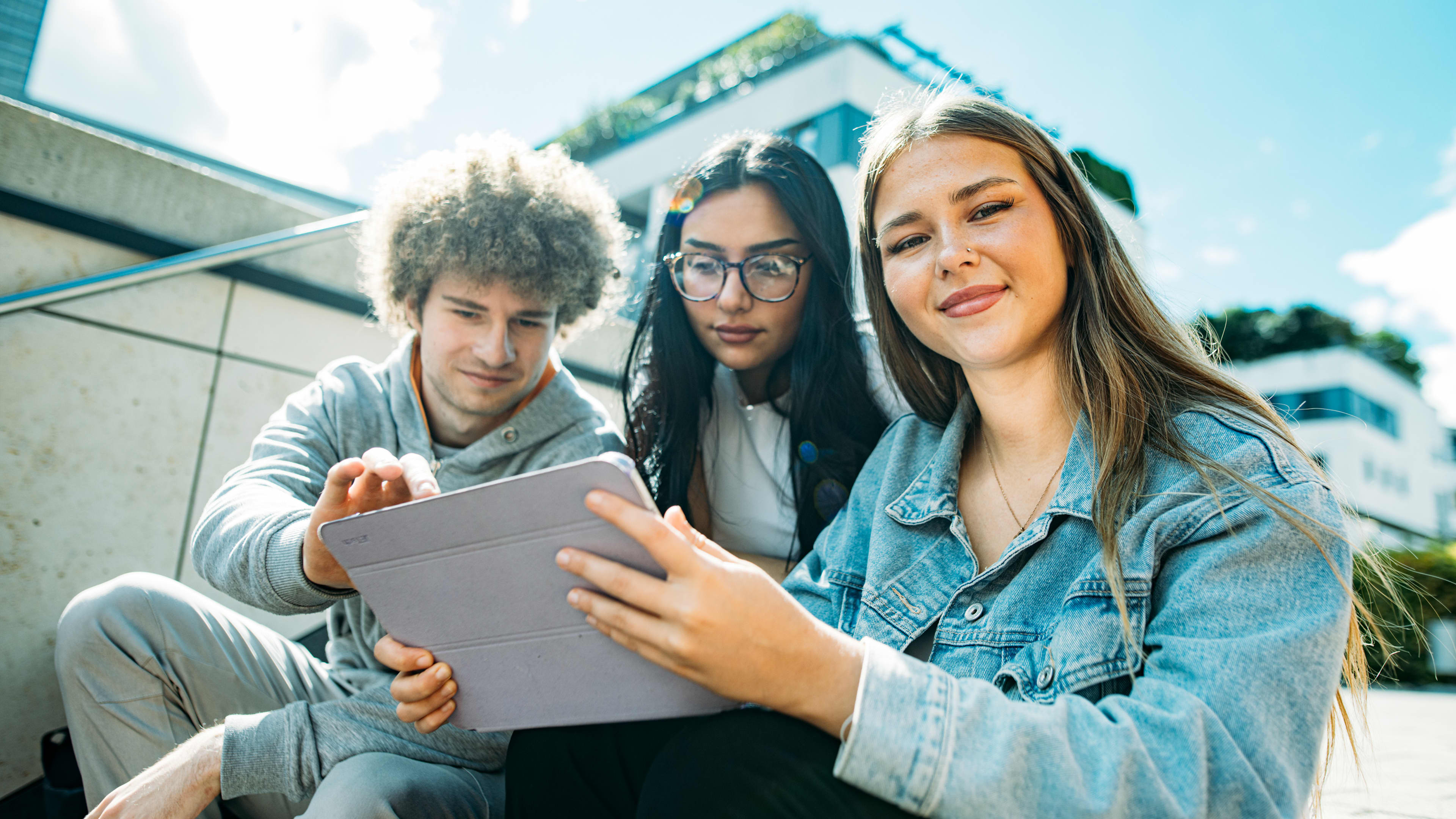 Three university students sitting outdoors on campus, collaborating and studying together using a tablet in front of a modern academic building in Germany.