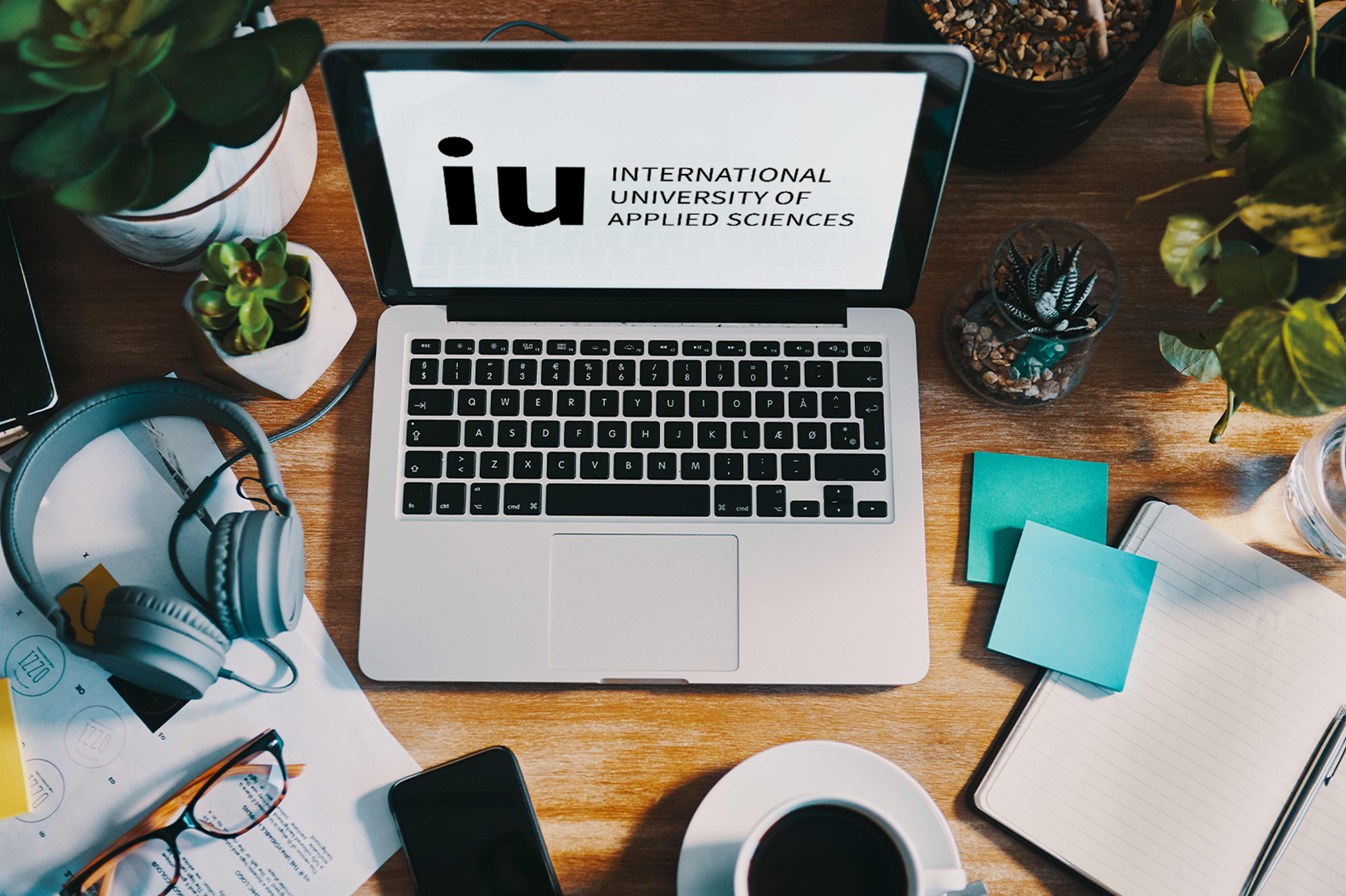 A laptop on a wooden desk displaying the IU University logo, surrounded by plants, headphones, a notebook, and a cup of coffee, illustrating a student studying in a home setup.