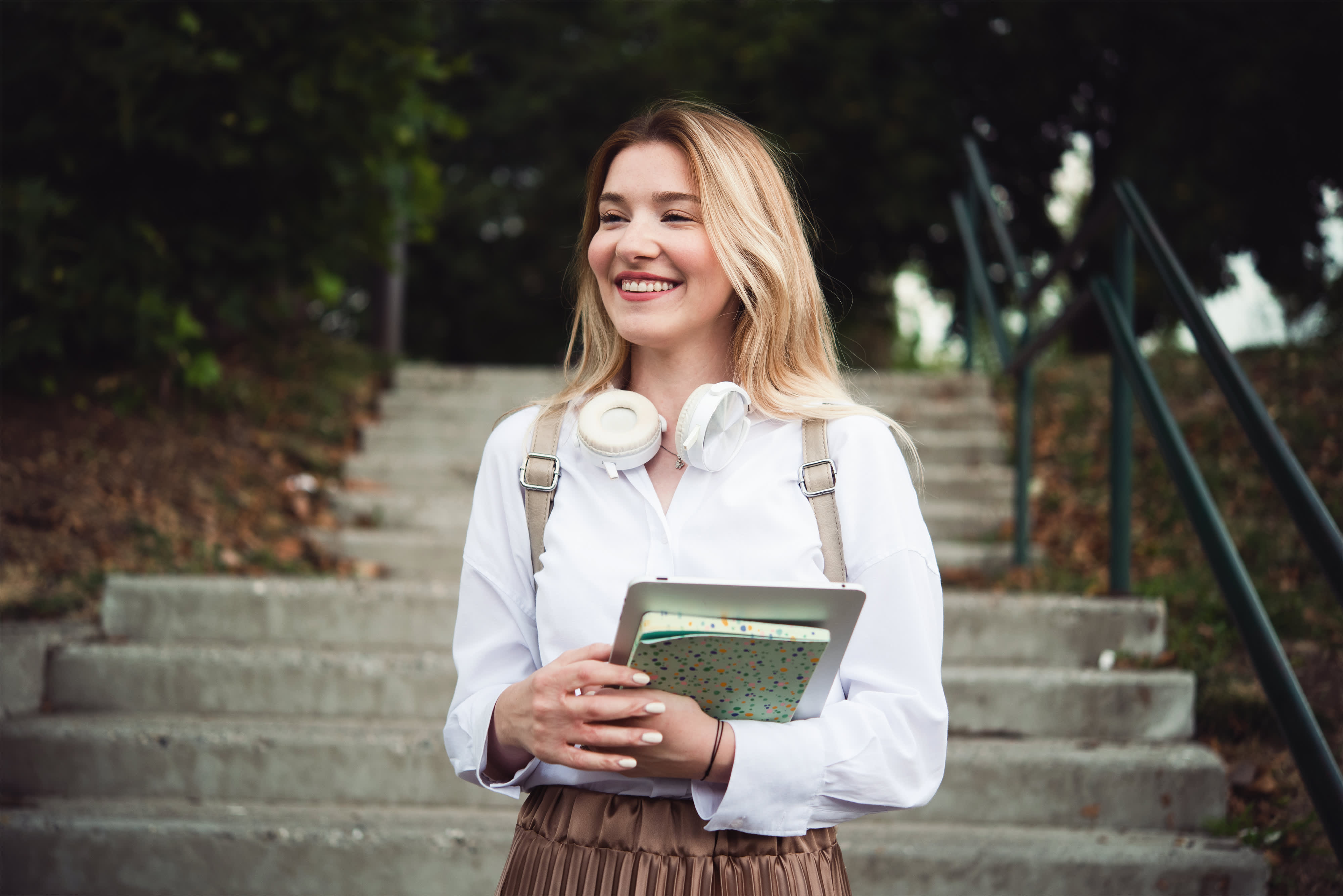 Titelbild der Infoseite "Duales Fernstudium": Duale Studentin mit langen blonden Haaren steht auf einer Treppe, hat einen Rucksack auf dem Rücken und hält Laptop sowie Notizbuch in der Hand