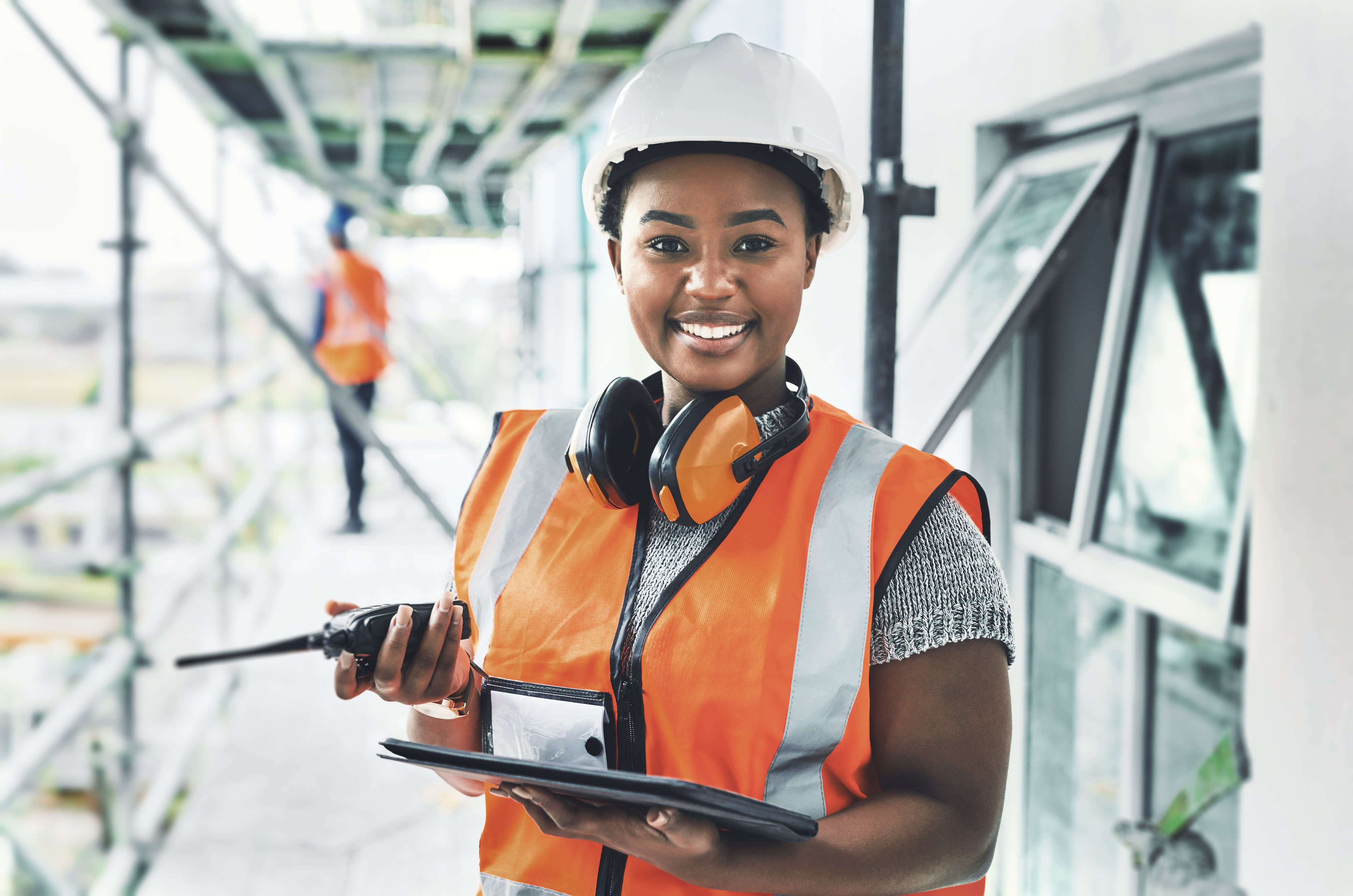 Titelbild für Infoseite “duales Studium Bauingenieurwesen”: Lachende duale Studentin mit Sicherheitsweste, Helm und Funkgerät auf einer Baustelle