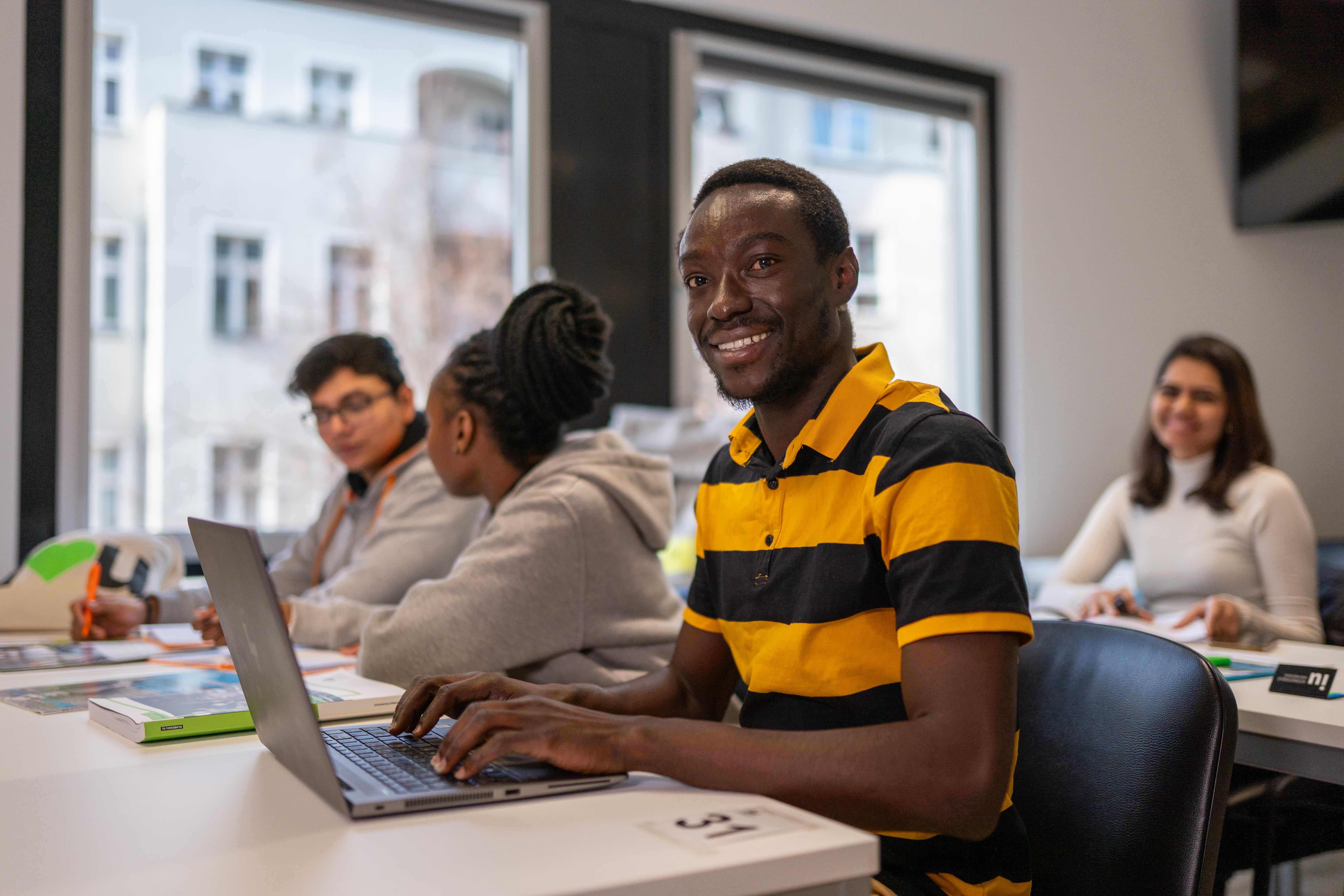 A student uses his laptop and smiles during class, while other students engage in conversations.