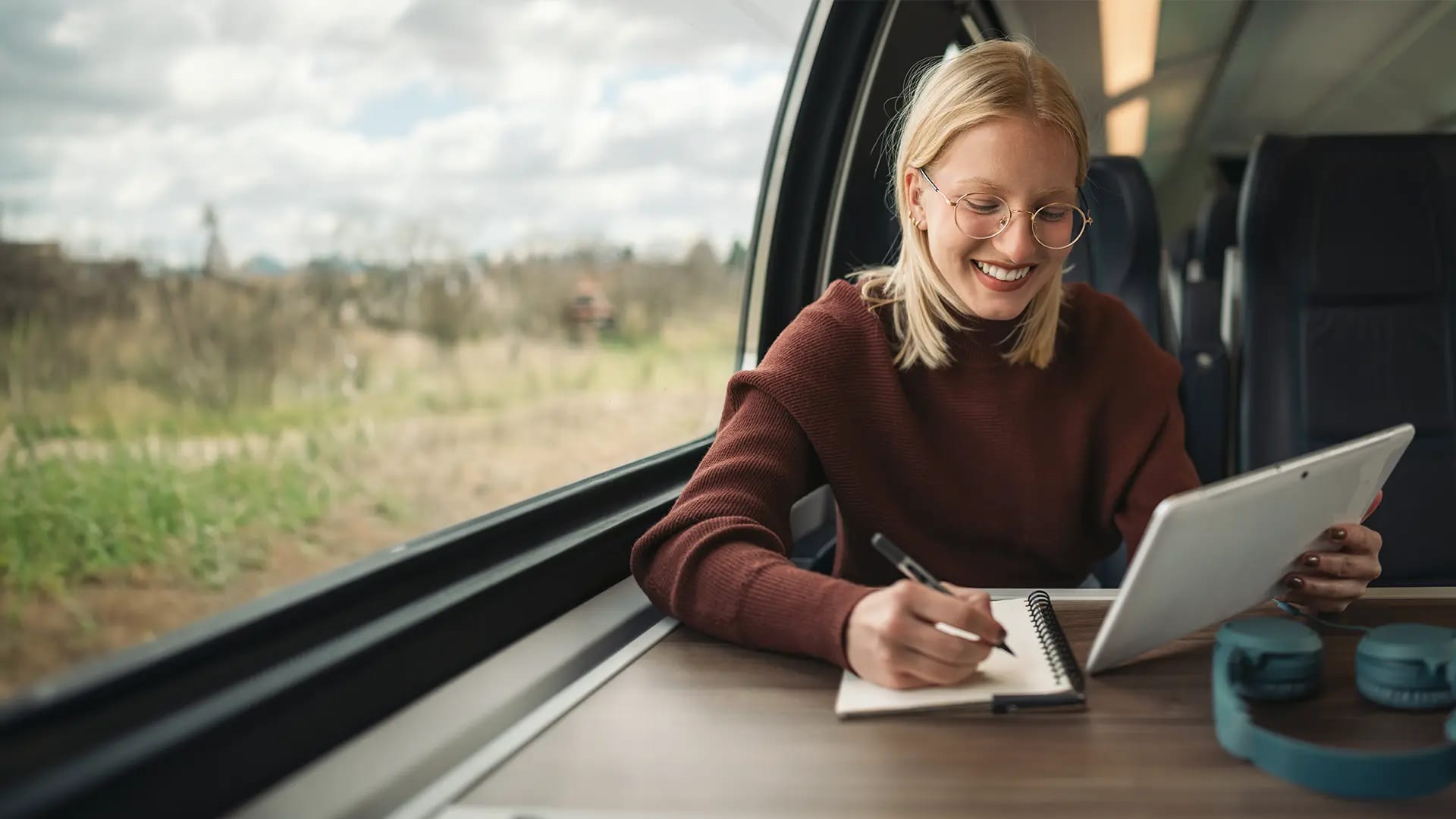 Titelbild für Artikel zum Teilzeitstudium: Lächelnde Studentin sitzt mit Tablet und Notizbuch in der Bahn