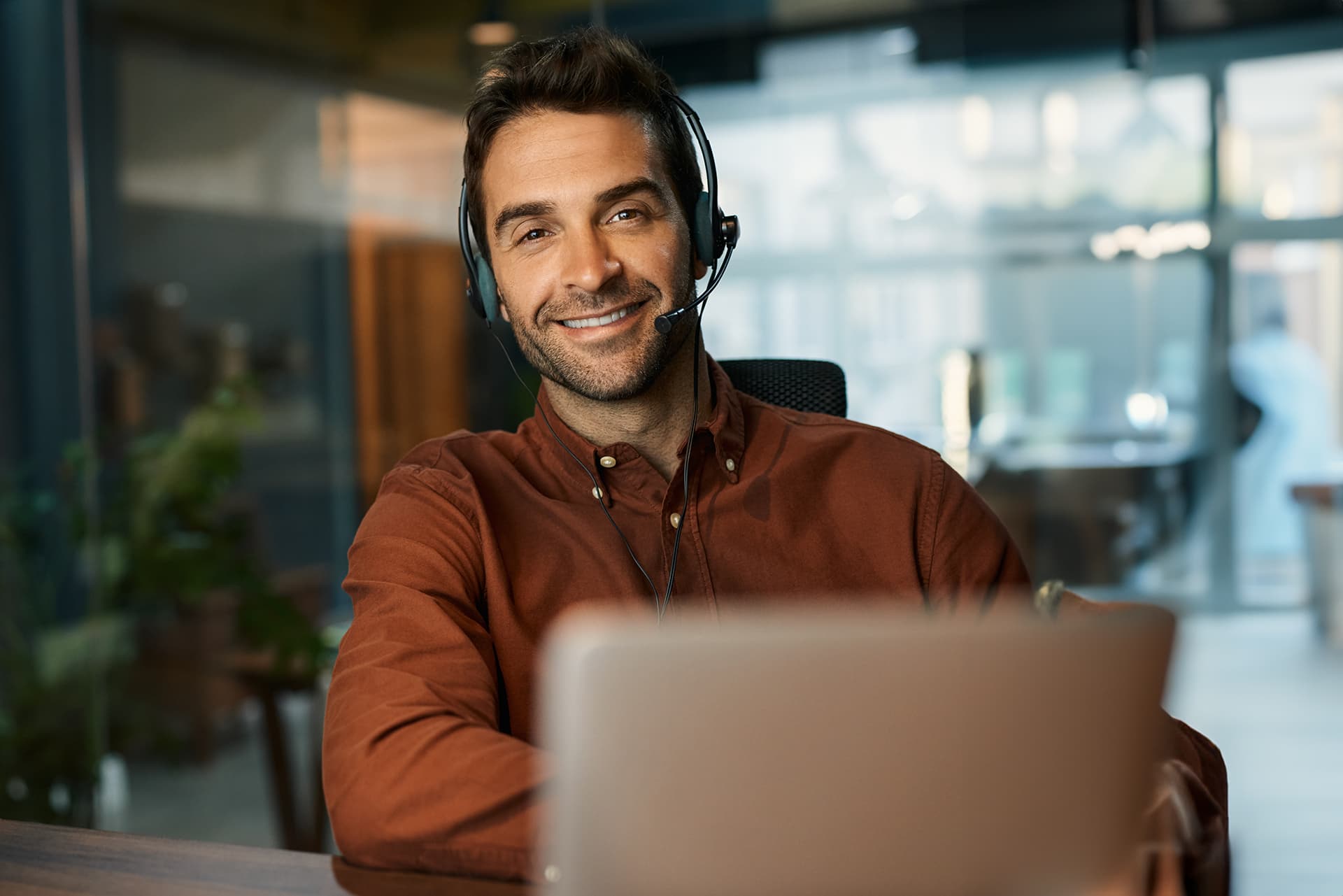 A cheerful man in a rust-coloured shirt working on his laptop, as part of a business degree that's an online MBA program, teaching strategic management skills and business administration.