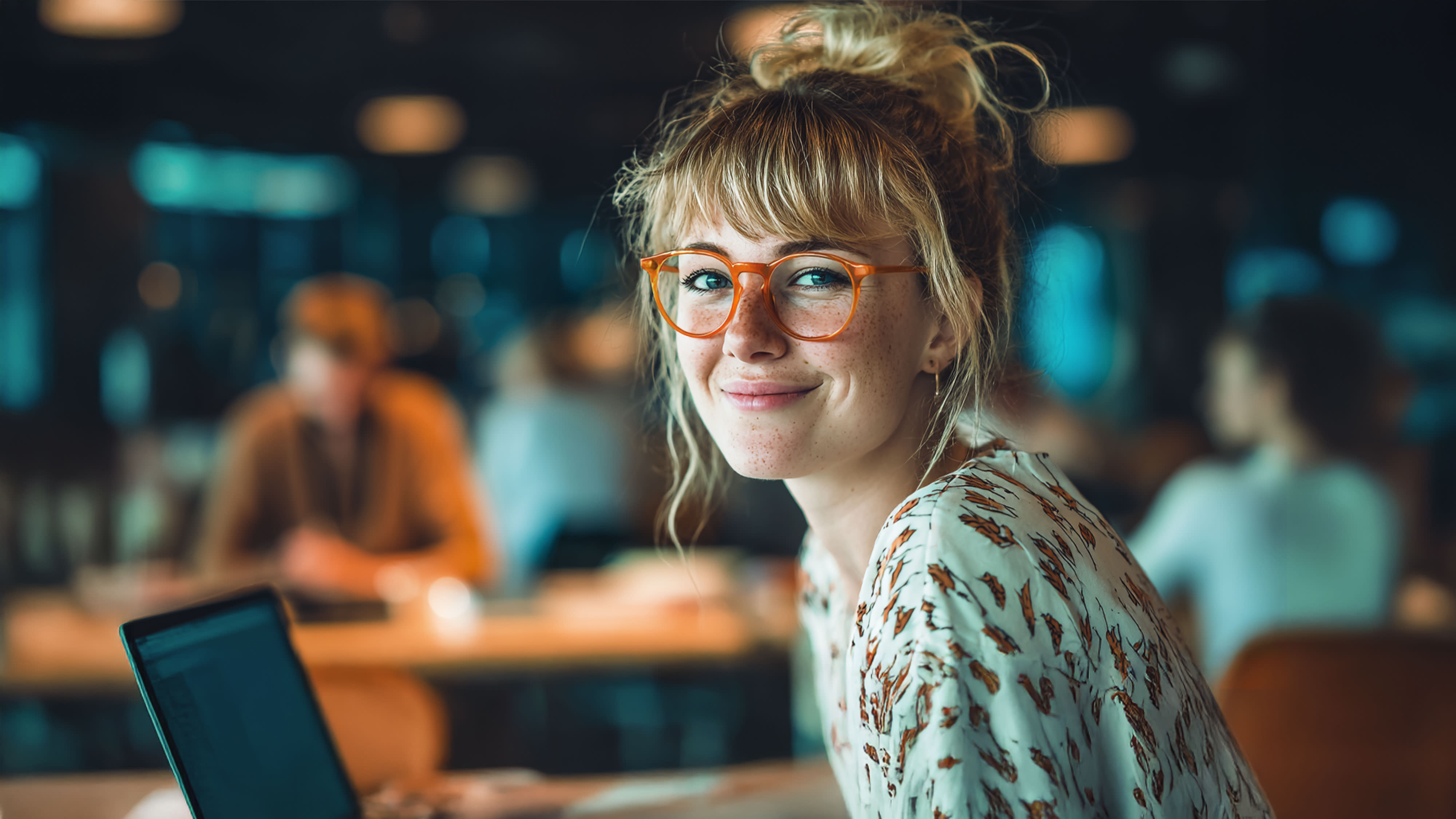 Junge Frau mit Brille und Dutt lächelt in die Kamera während sie an einem Laptop in einem modernen Büro oder Coworking-Space arbeitet, mit unscharfen Personen im Hintergrund