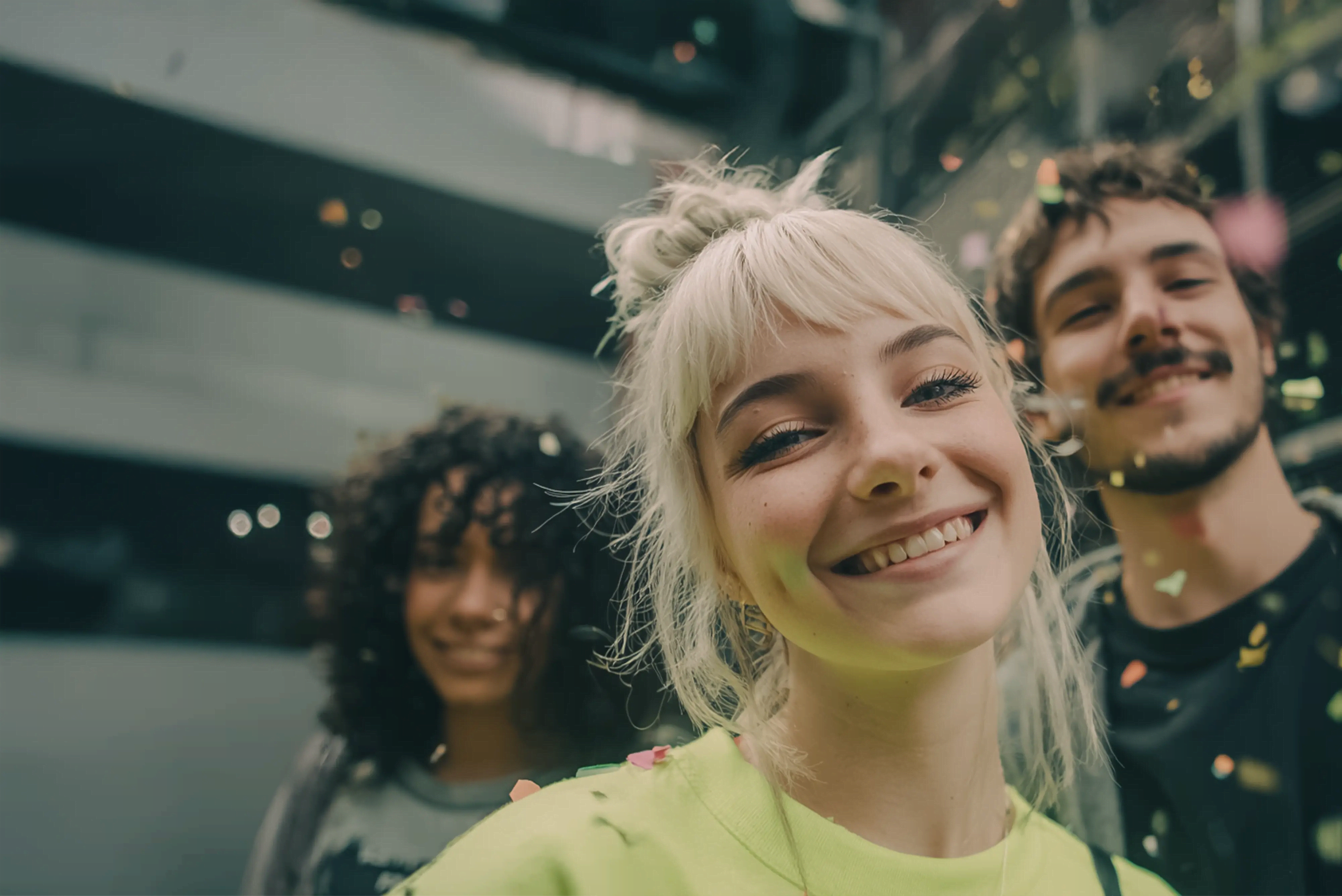 Drei Studierende lachen in die Kamera, Konfetti fliegt; unscharfer, urbaner Campus im Hintergrund
