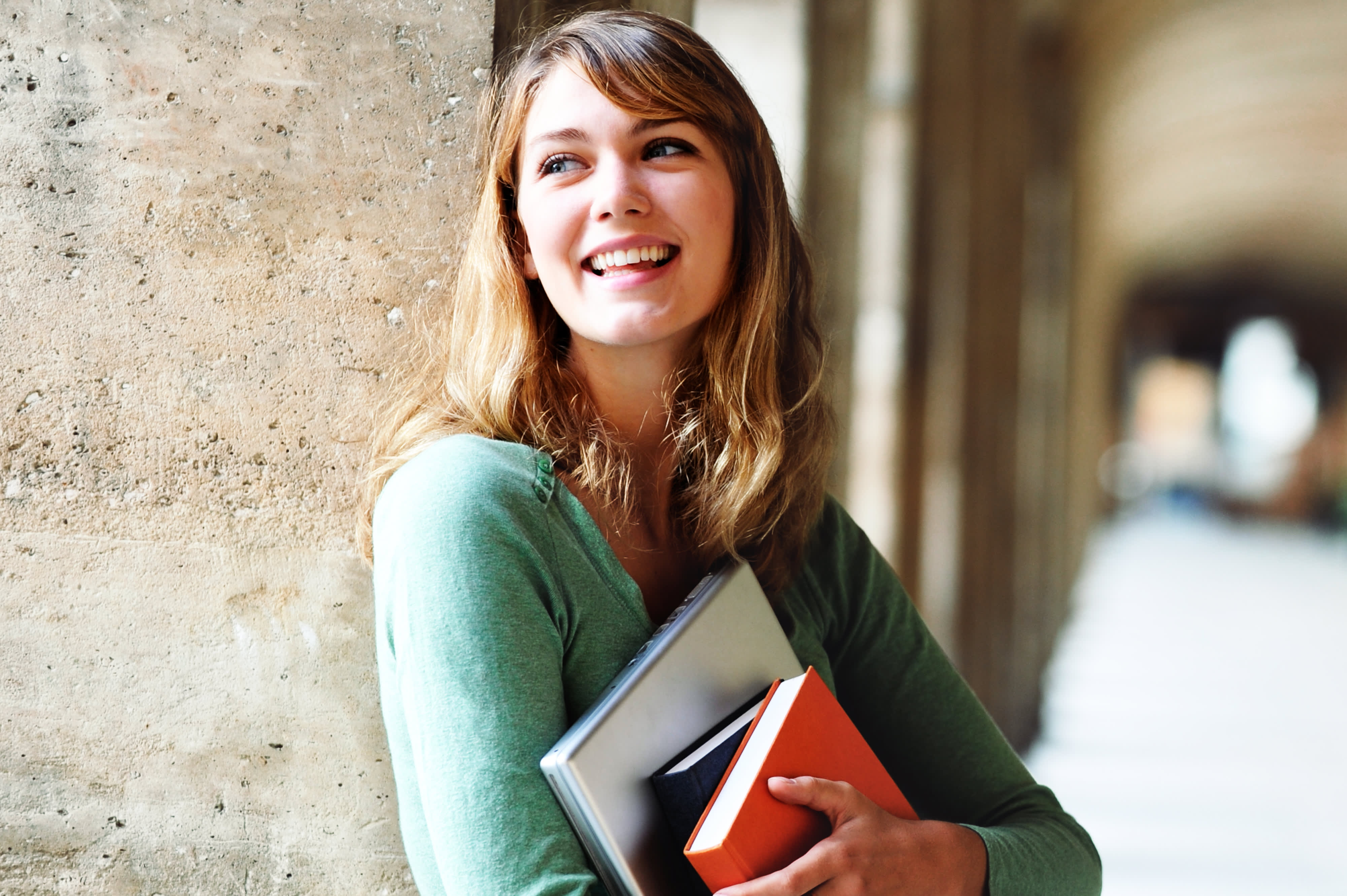 Titelbild der Infoseite "Ausbildung an der IU": Auszubildende lehnt an einer Säule und hat Bücher und Laptop im Arm