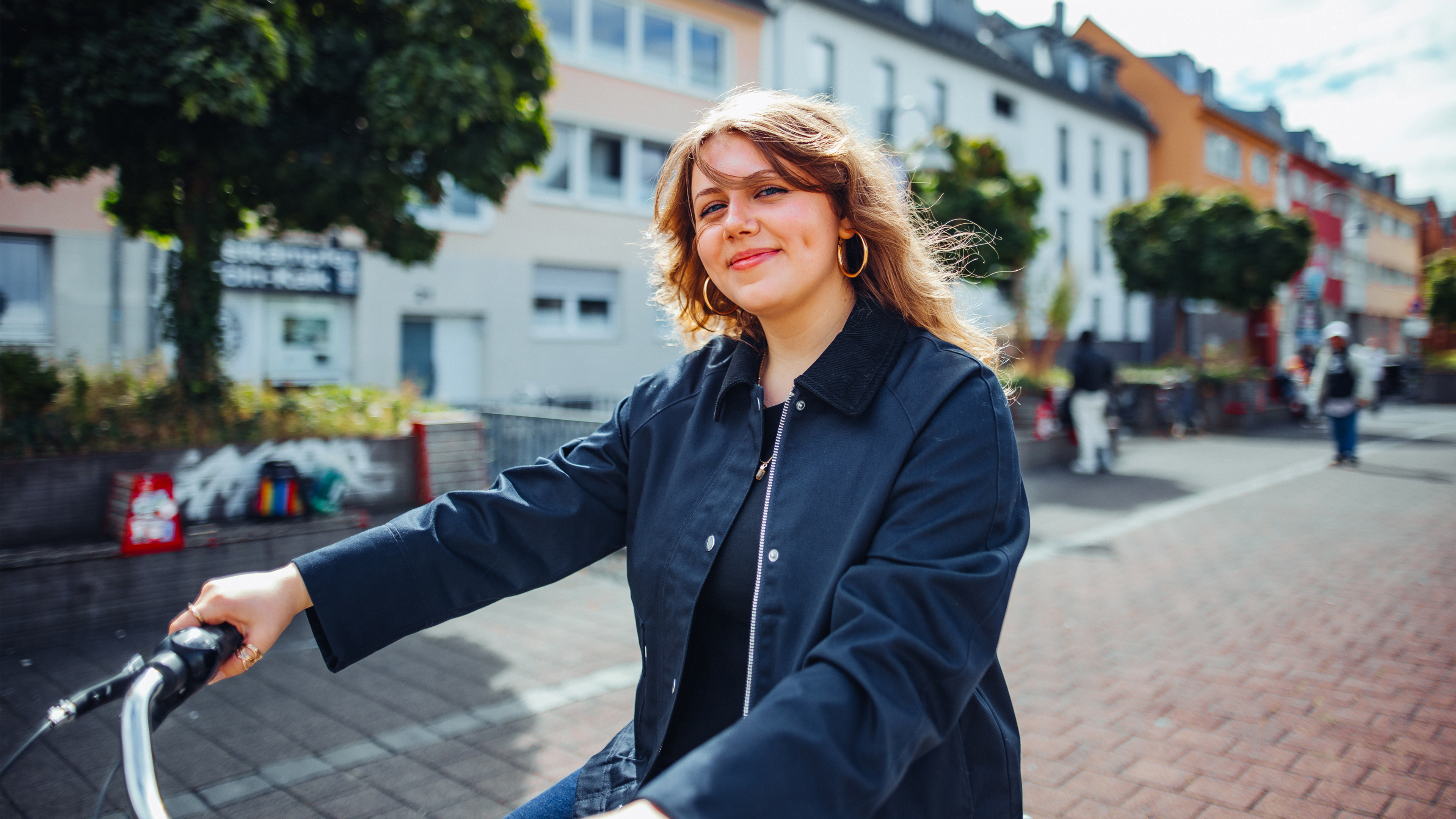 Young woman with curly hair, dimples, and hoop earrings smiles while riding a bicycle on a brick path in front of colorful buildings and trees.