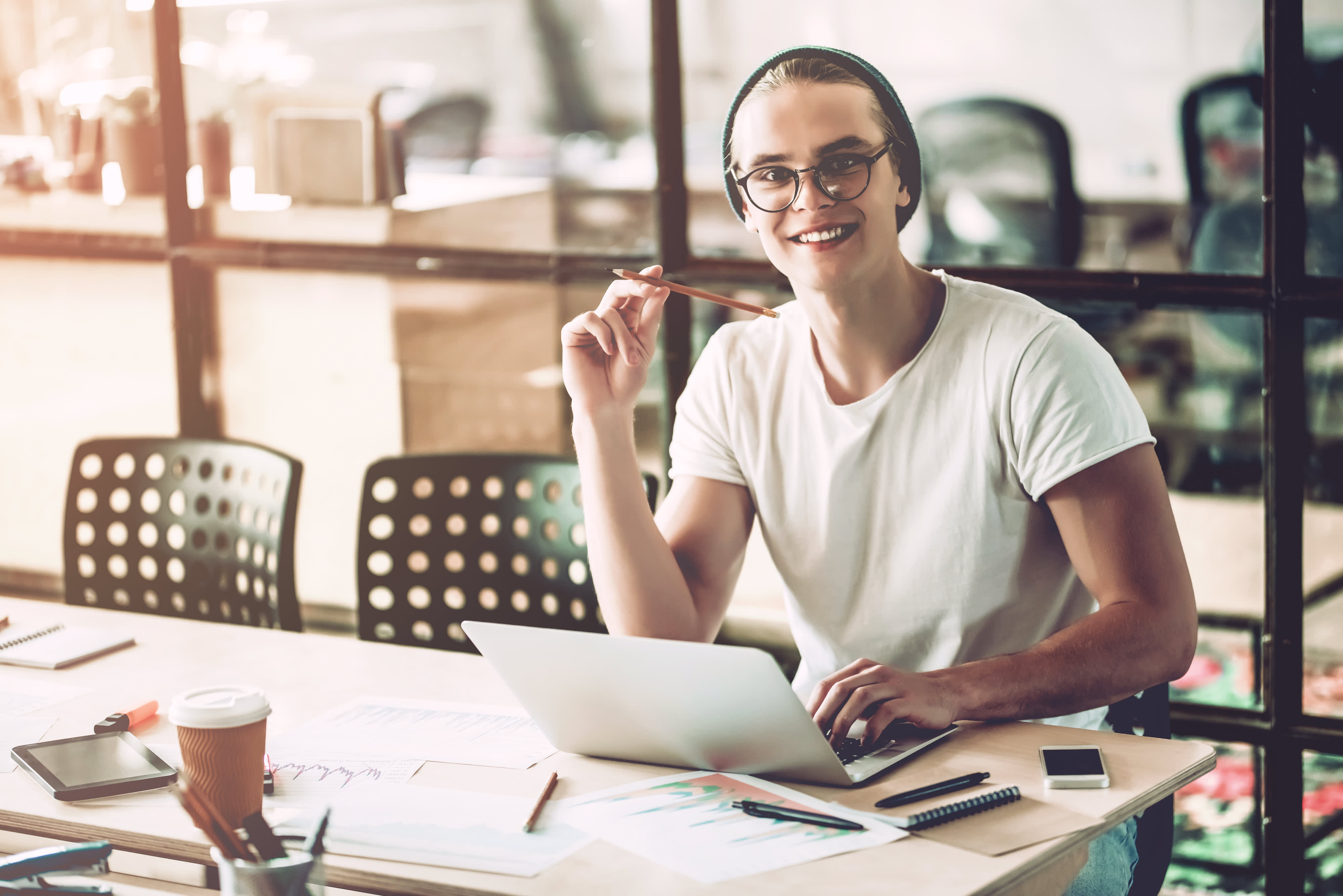 Titelbild für Infoseite “duales Studium Marketingmanagement": Lächelnder dualer Student hält einen Bleistift in der Hand und sitzt in einem Büro am Schreibtisch mit Laptop vor sich