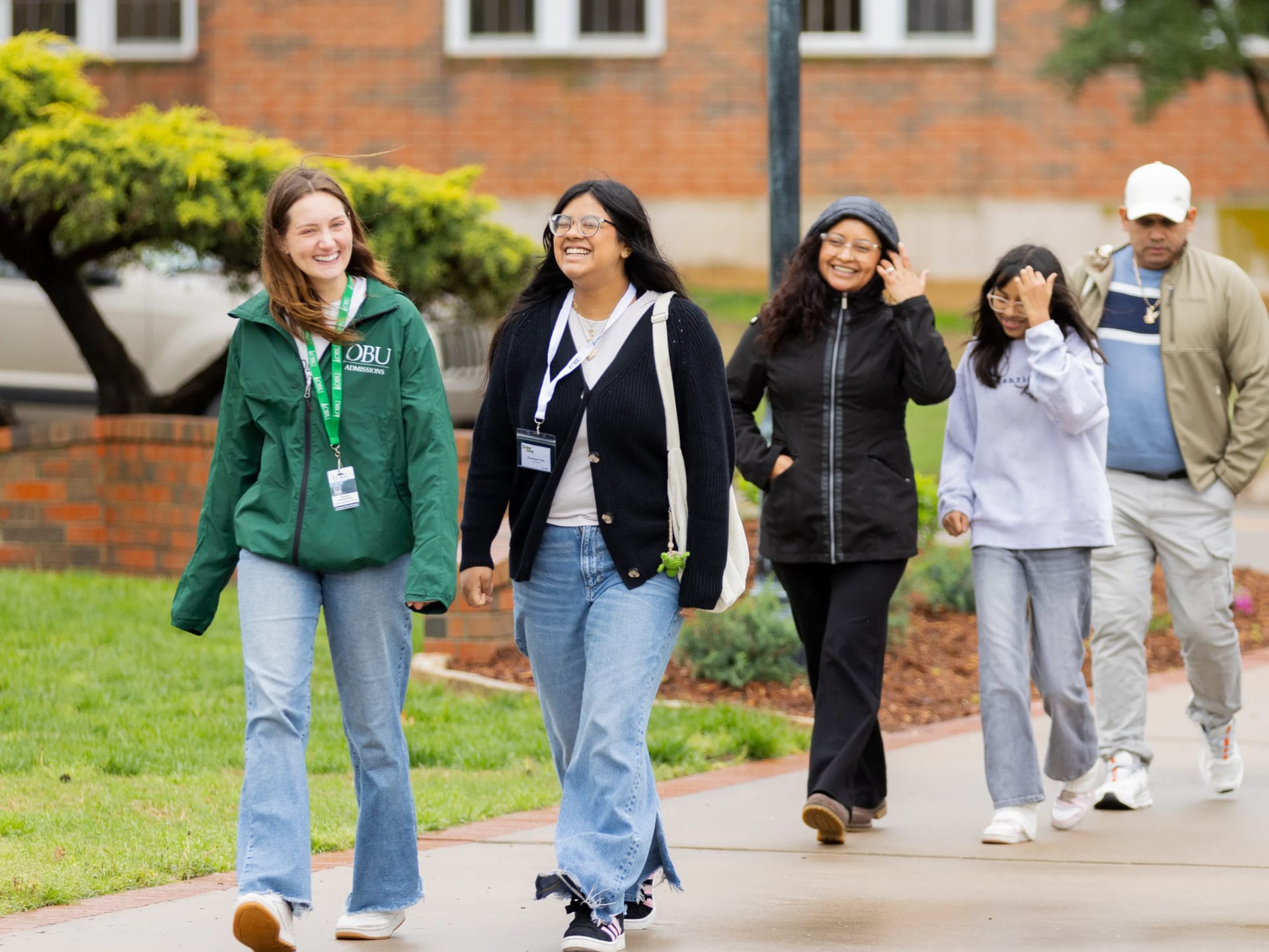 A group of OBU students and staff walk together and laugh on a campus sidewalk in front of a brick building.