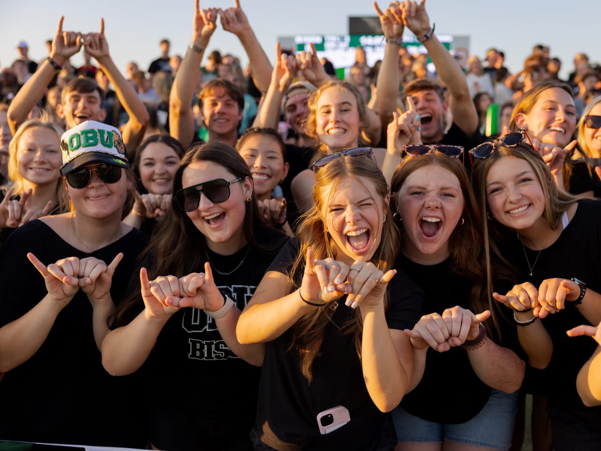 A large crowd of cheering OBU students throw up hand signs and pose for the camera at an outdoor event.