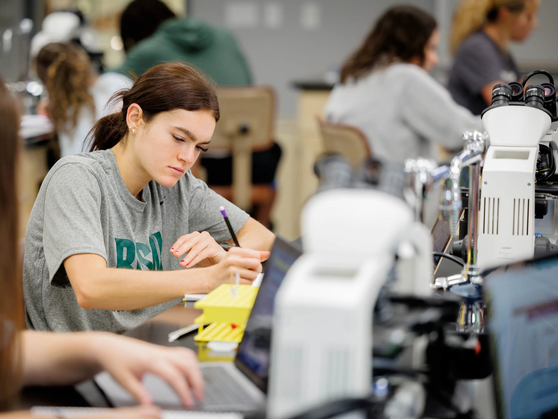A student writes in a notebook while working at a lab bench surrounded by microscopes and other students.