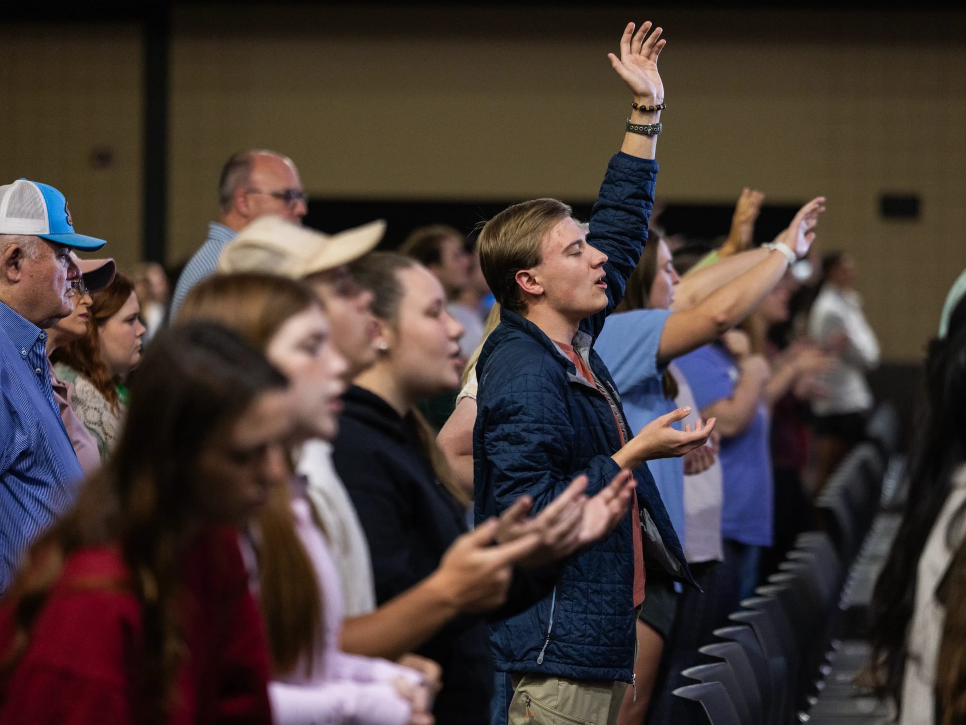 A young man raises his hand in worship during a chapel service, surrounded by other students and attendees with hands raised or clapping.