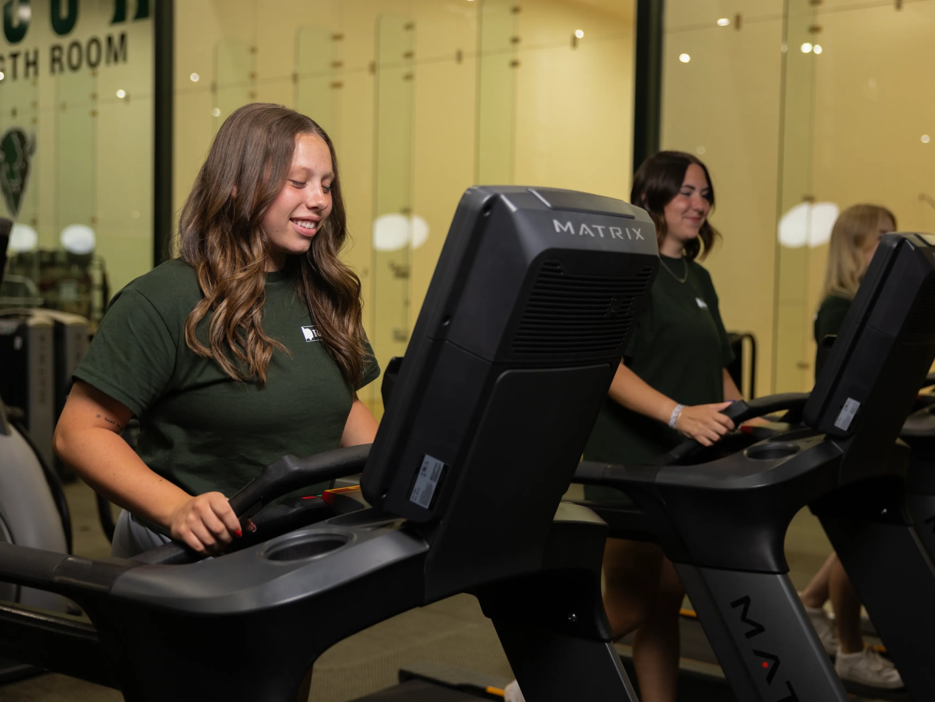 Two students smile while working out on treadmills in a campus fitness room.