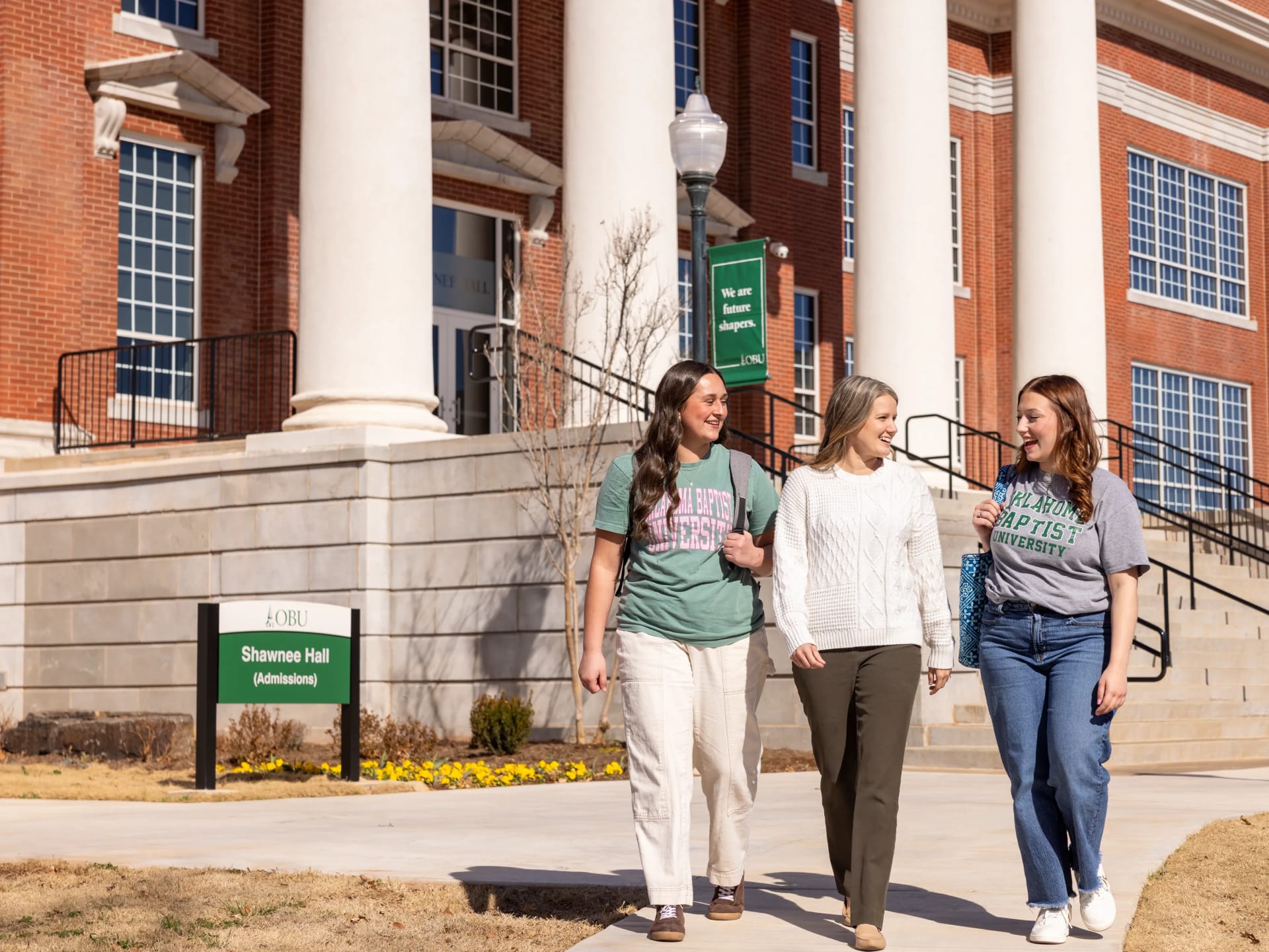 Three smiling women in Oklahoma Baptist University t-shirts and a cream sweater walk together on a sidewalk in front of Shawnee Hall, past a sign reading "Shawnee Hall (Admissions)."