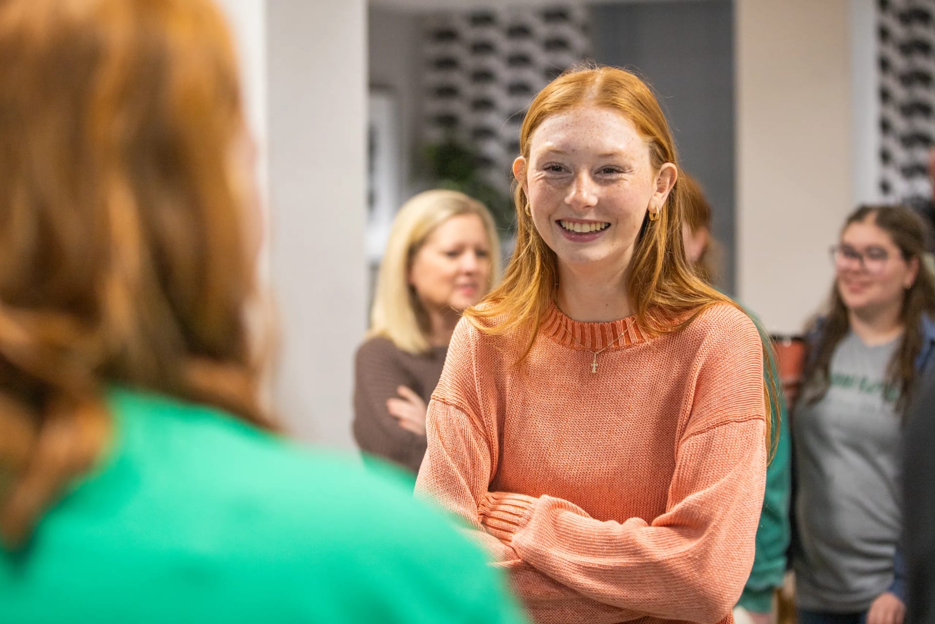 A smiling young woman with red hair and freckles, wearing an orange sweater and a small cross necklace, stands with her arms crossed during a campus visit, with other visitors blurred in the background.