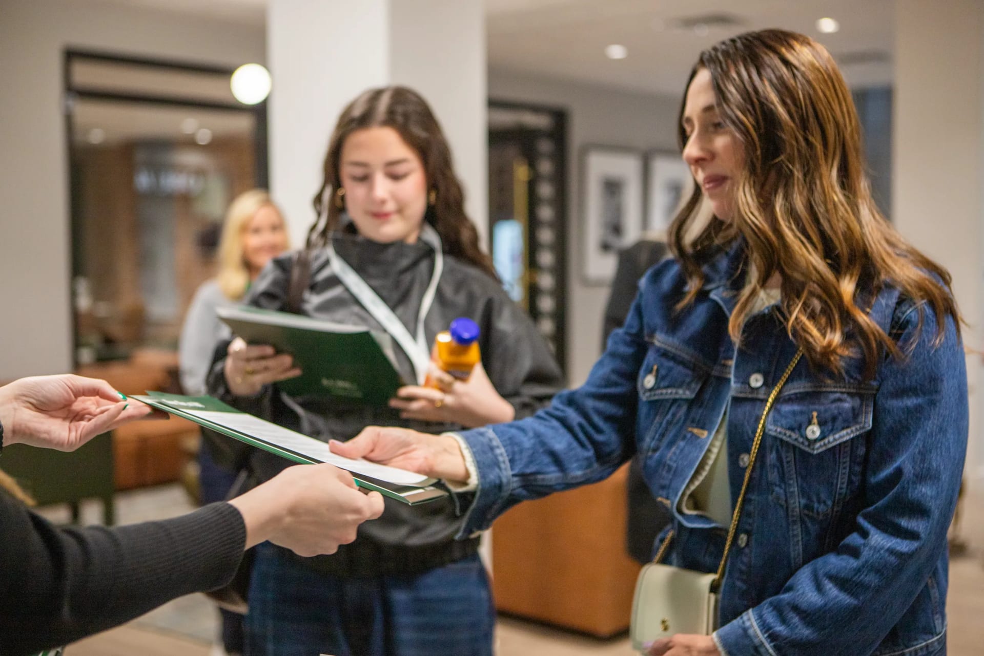 A staff member in a denim jacket hands a green folder of materials to another woman, while a student and another attendee look on nearby.