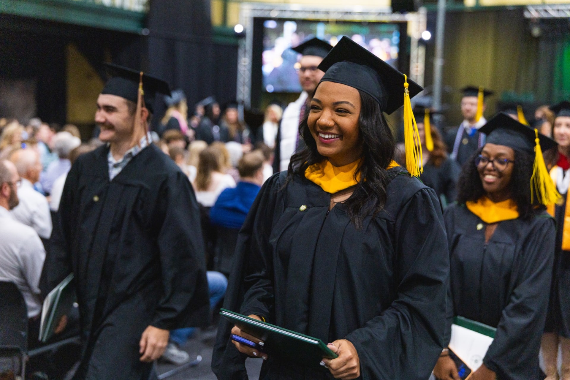 A student in a black graduation gown and cap with a gold honor cord and tassel smiles while walking through the crowd, holding her diploma cover, with fellow graduates beside her.