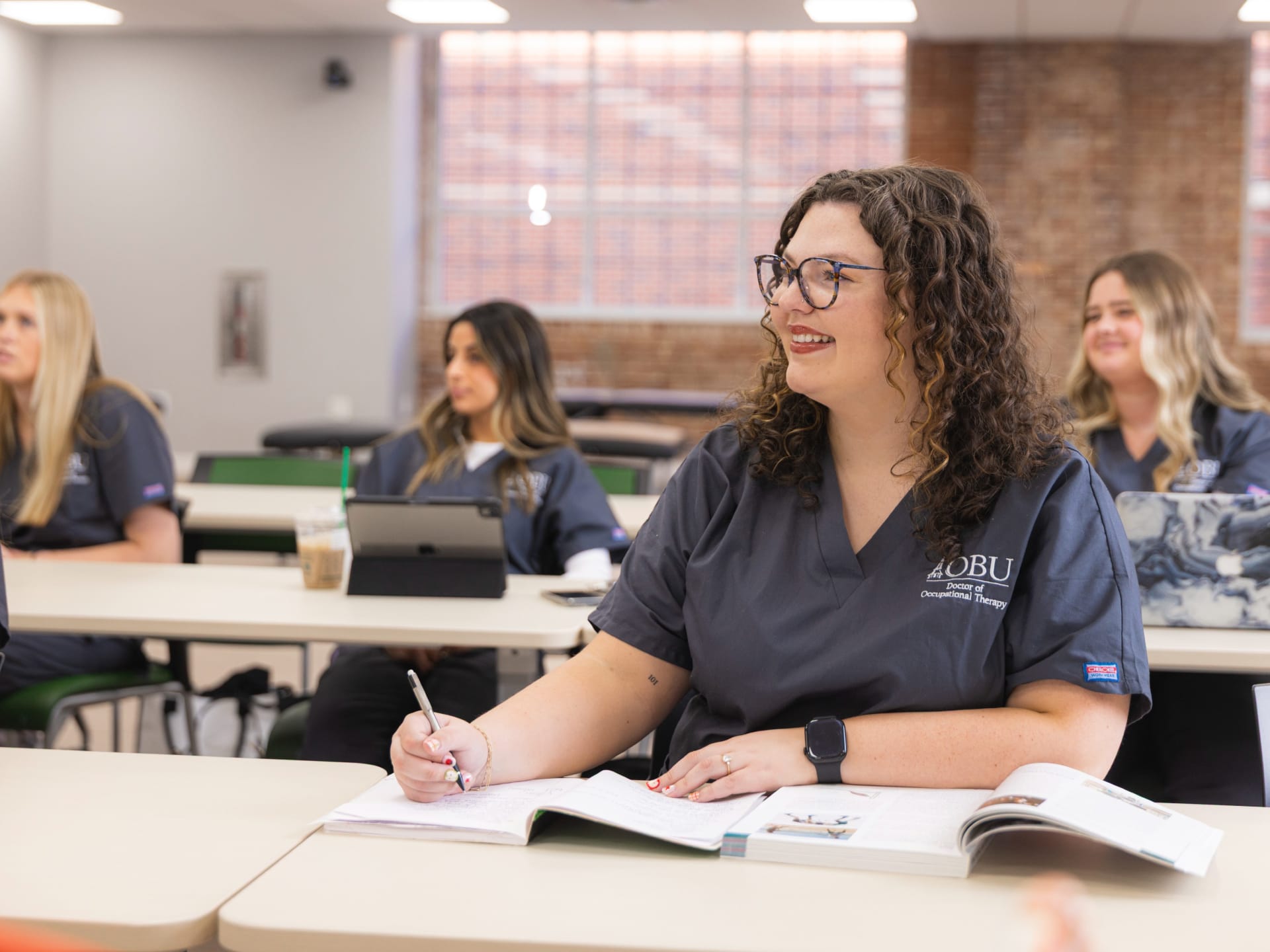 A student wearing an OBU Doctor of Occupational Therapy scrub top smiles while taking notes in class, with classmates working at tables in the background.