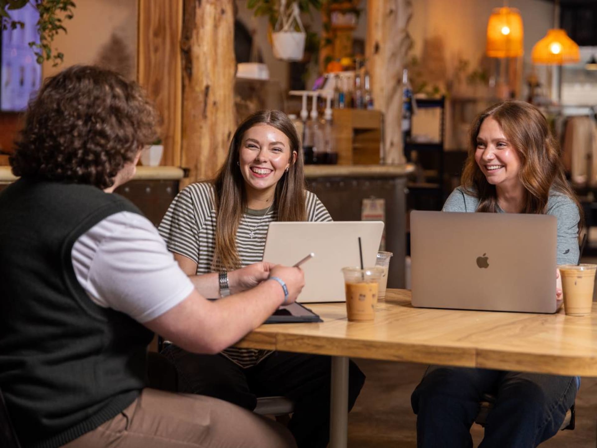 Three people sit and stand around a wooden table at a coffee shop, two women seated with laptops open and smiling at a man across from them, warm string lights and wood beams in the background.