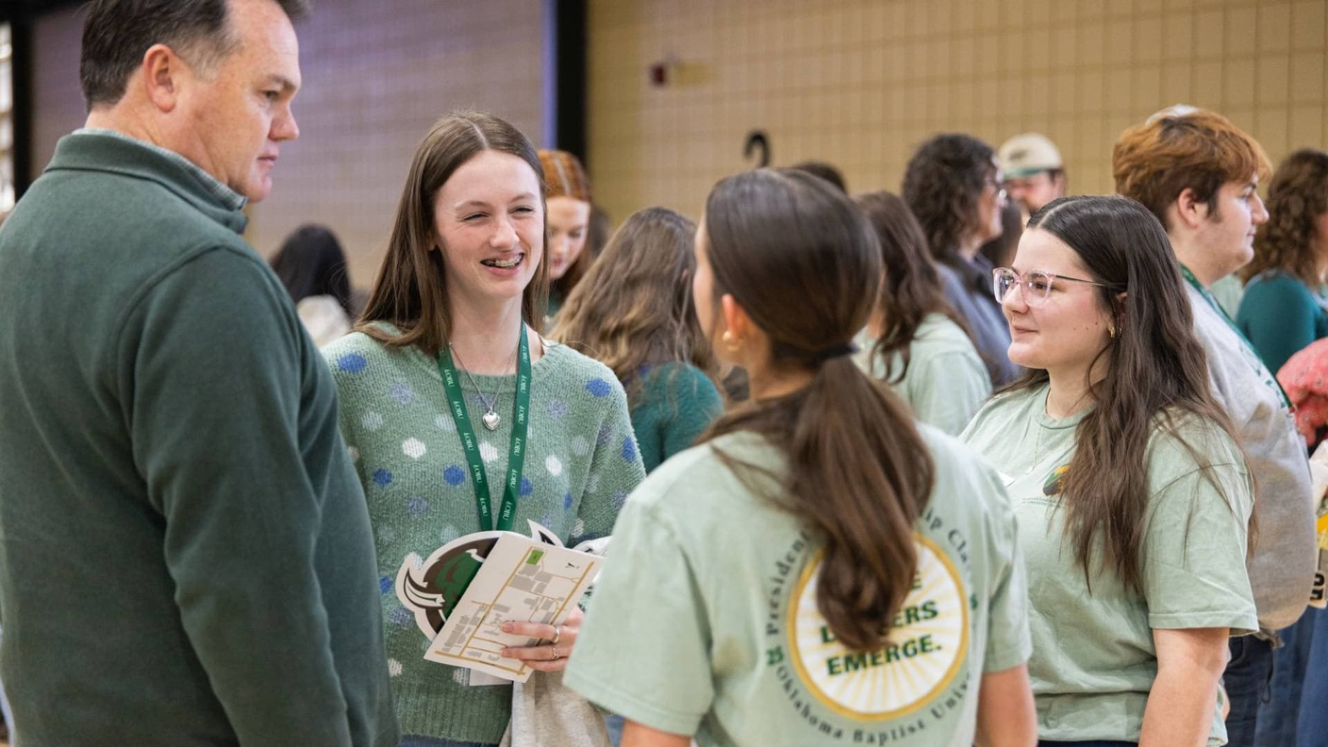 Students in light green President's Leadership Class T-shirts talk with a man in a dark green pullover inside a campus gymnasium. A smiling student wearing an OBU lanyard holds a campus map as the group chats.