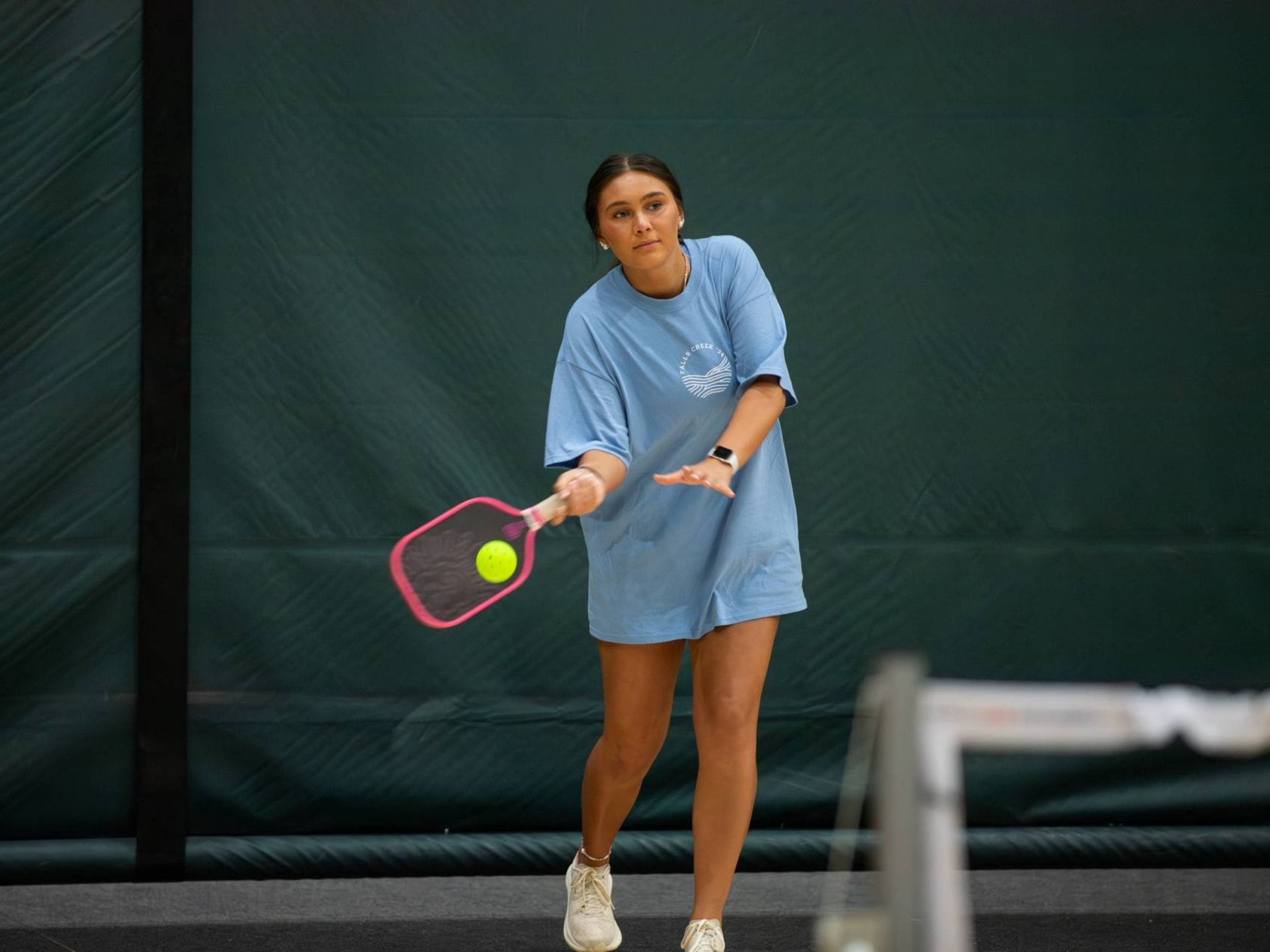 A woman in an oversized light blue T-shirt swings a pink pickleball paddle at a yellow ball during a game on an indoor court.