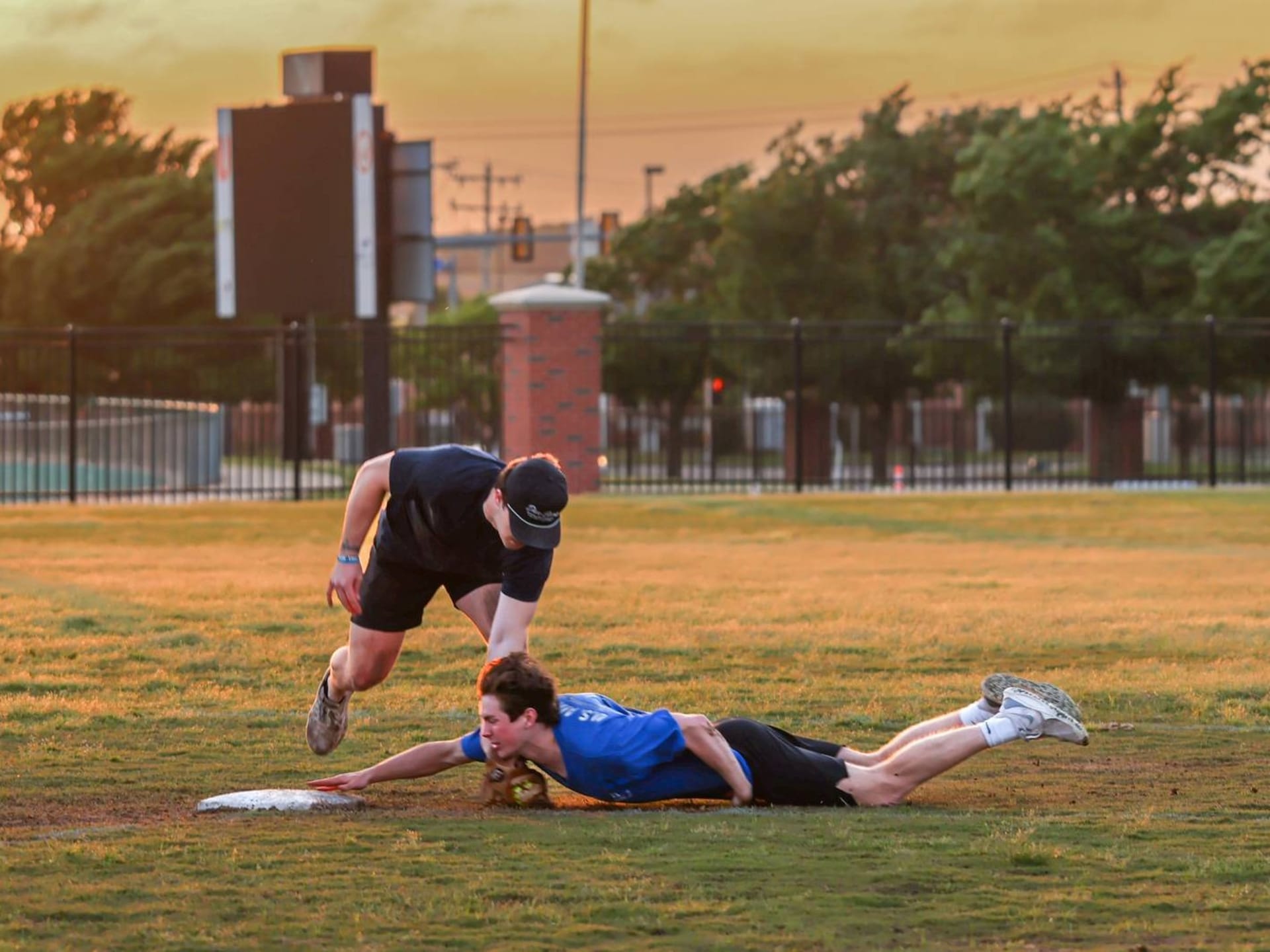 A player in a blue shirt dives headfirst toward a base at sunset, hand outstretched to touch it, as a player in a dark shirt and cap leans over him with a glove to make the tag.