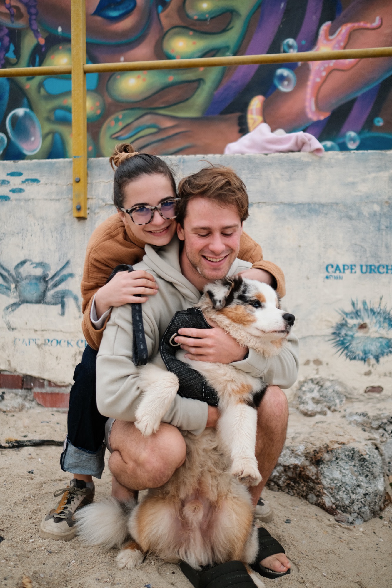 Tristan and Erin holding a puppy by a painted beach wall