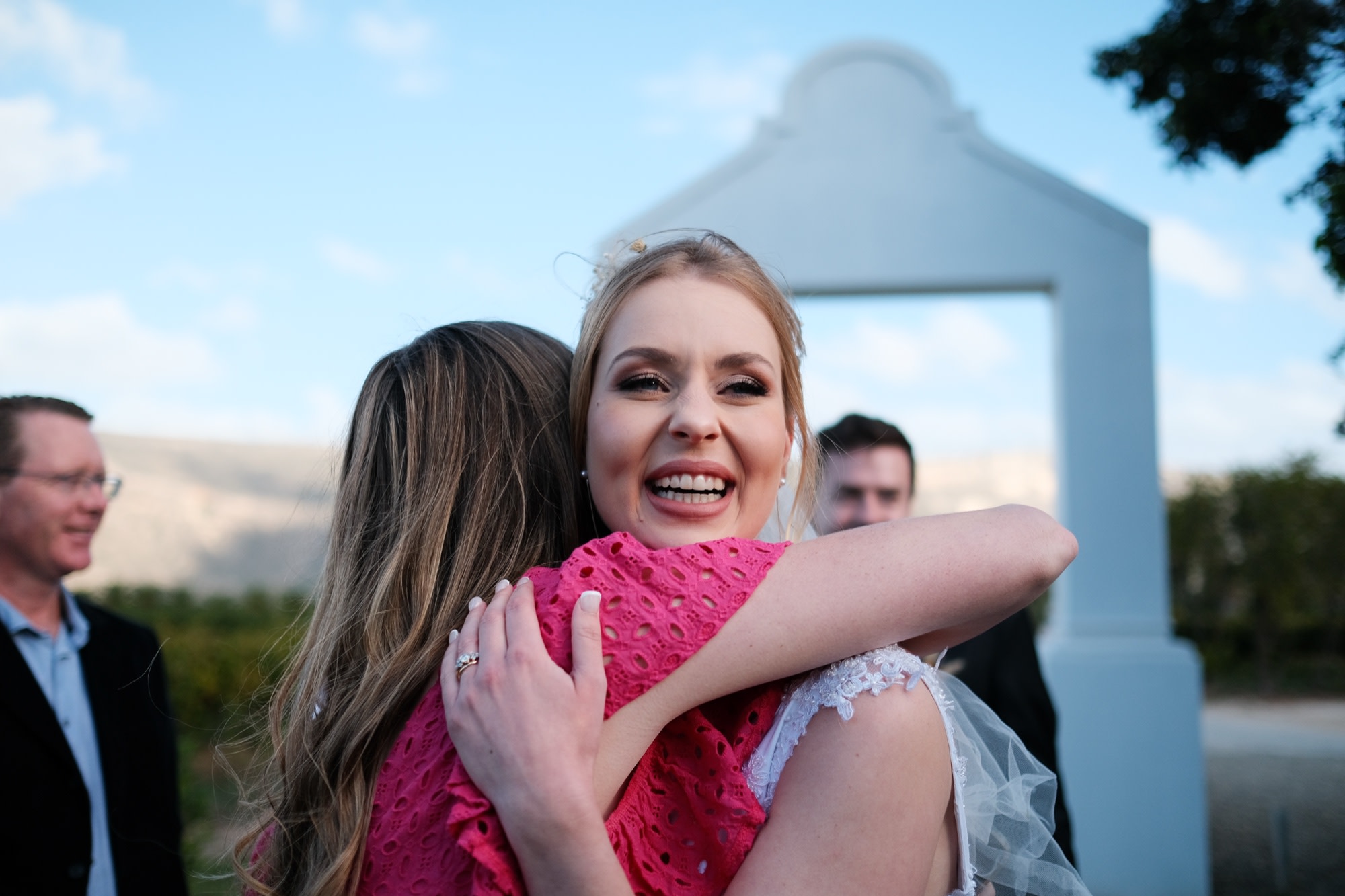 The bride hugging a guest, both beaming, by the white arch
