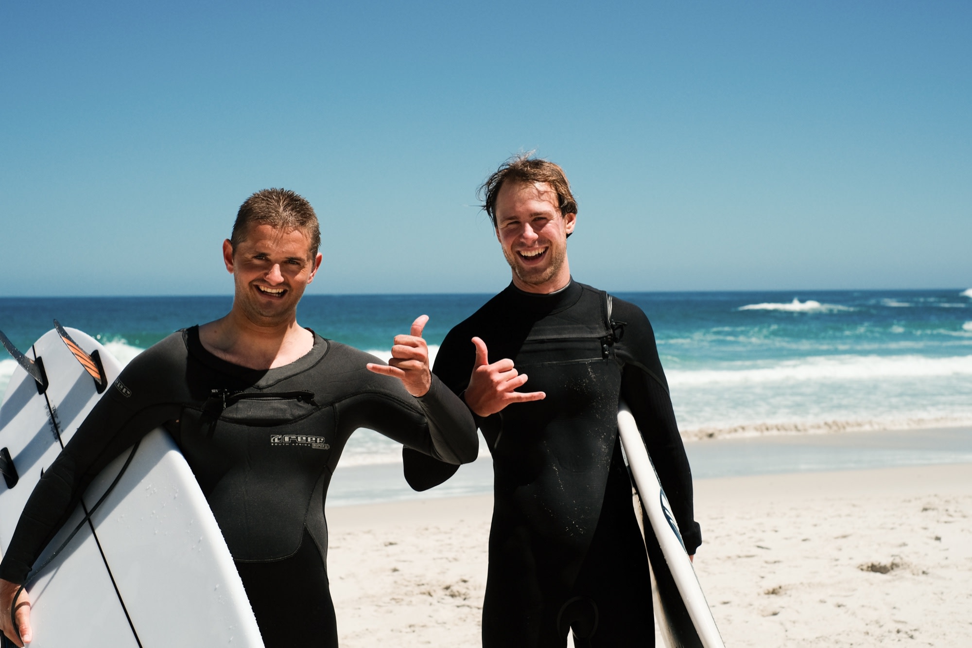 Two friends in wetsuits throwing shakas on the beach, boards in hand