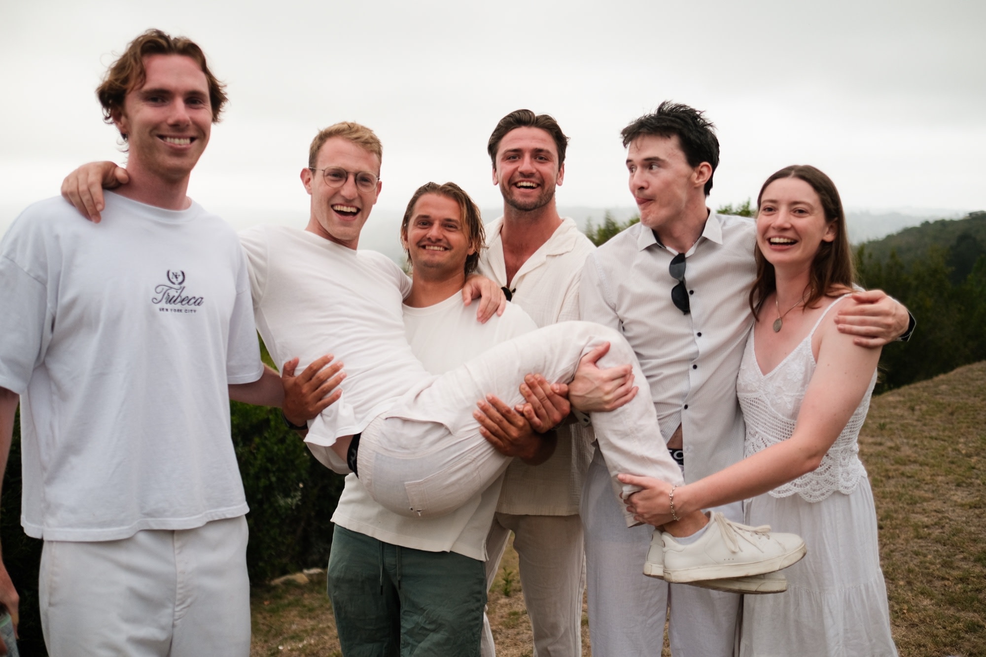 Six friends in white, laughing, carrying one of them aloft