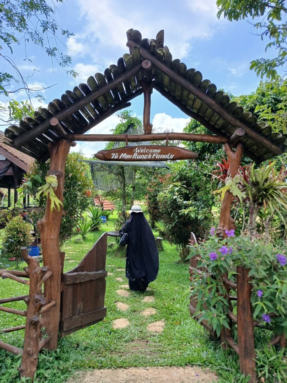 Gerbang kayu dengan papan bertuliskan ‘Welcome To Mini Ranch Family’; seseorang berjalan menjauh dengan punggung menghadap kamera.