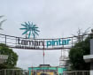 Gerbang masuk Taman Pintar Yogyakarta berbentuk lengkung dengan tulisan taman pintar, dengan bilik pengunjung kosong di sisi kiri dan kanan, pagar di bagian depan, langit mendung, dan poster protokol kesehatan kecil di bilik kanan.