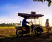 Suasana senja di jalan kampung saat musim mudik: warga membawa barang bawaan berpapasan dengan kendaraan bermotor kecil di jalan bata, latar sawah dan pohon tropis.