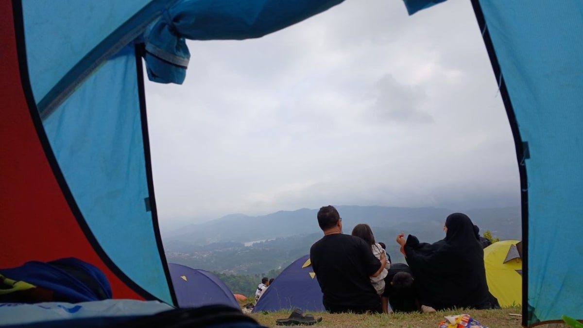Seorang pria, anak kecil, dan perempuan berhijab duduk di mulut tenda menghadap pemandangan lembah dengan badan air di kejauhan