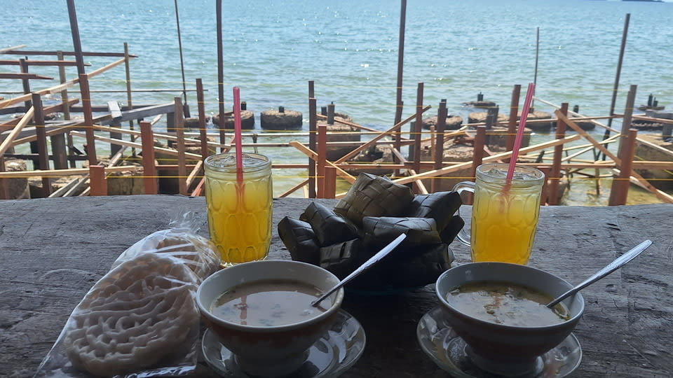Dua mangkuk coto Makassar berisi kuah berempah di atas meja kayu, ditemani ketupat berbalut daun pisang, dengan latar laut dan dermaga kayu.