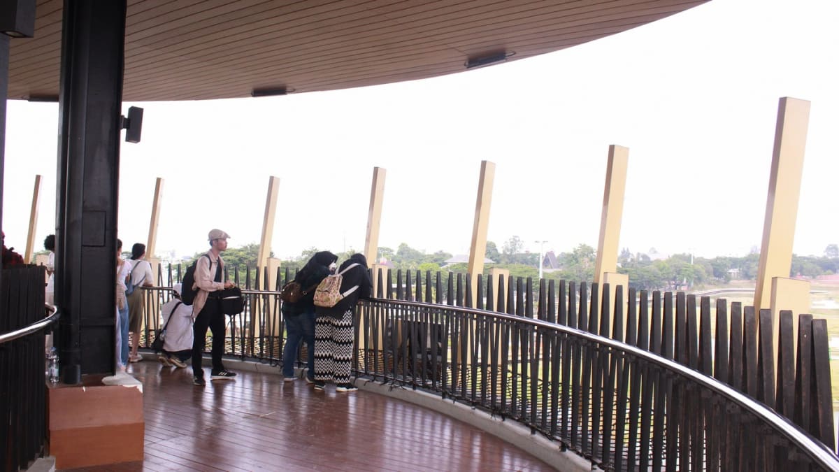 Pengunjung berdiri di balkon Menara Saudjana, Taman Mini Indonesia Indah, memandang telaga dan pepohonan hijau kawasan taman dari lantai atas.
