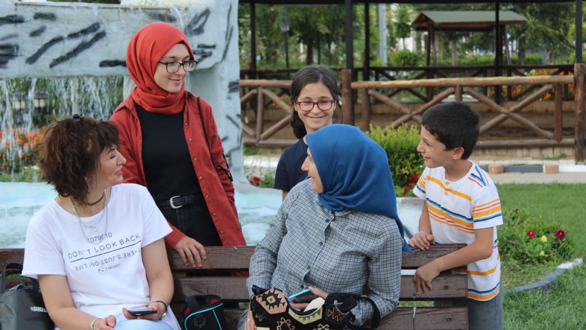 Seorang perempuan lansia, dua perempuan dewasa, dan dua anak berkumpul serta mengobrol di bangku taman dengan air mancur dan rumput hijau di latar.