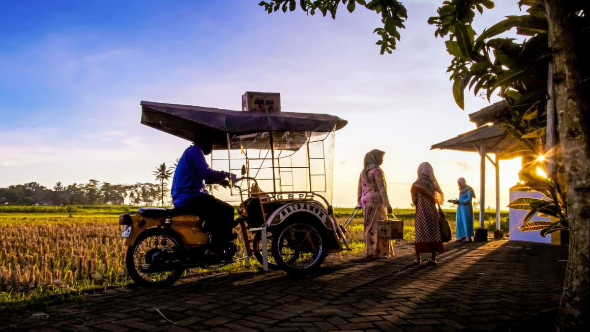 Suasana senja di jalan kampung saat musim mudik: warga membawa barang bawaan berpapasan dengan kendaraan bermotor kecil di jalan bata, latar sawah dan pohon tropis.
