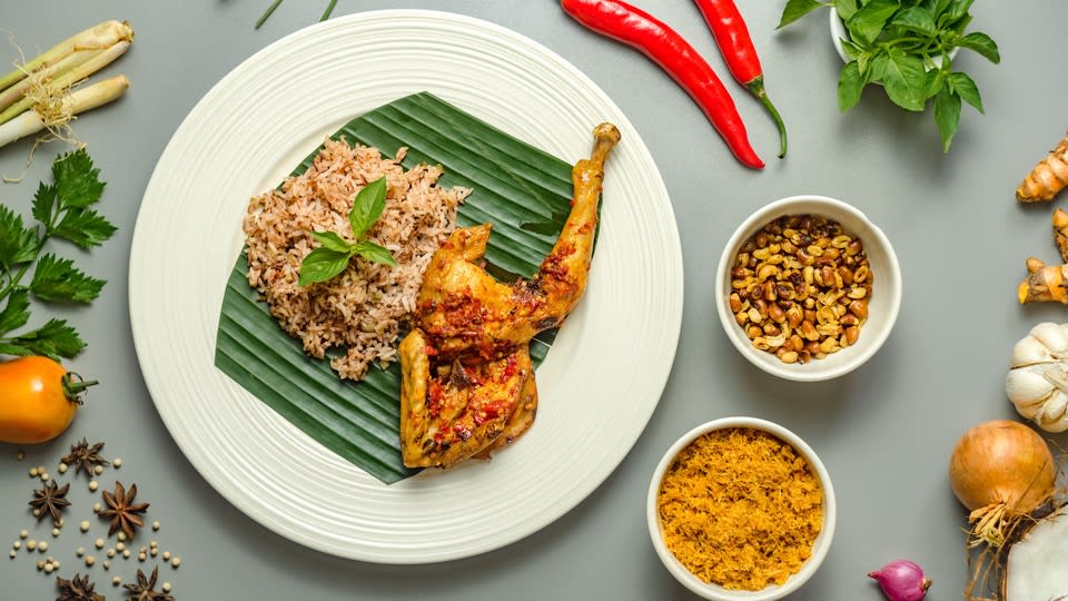 Hidangan ayam betutu berbumbu merah dengan nasi di atas piring beralas daun pisang, dikelilingi bahan bumbu seperti cabai, serai, bawang, dan kunyit.