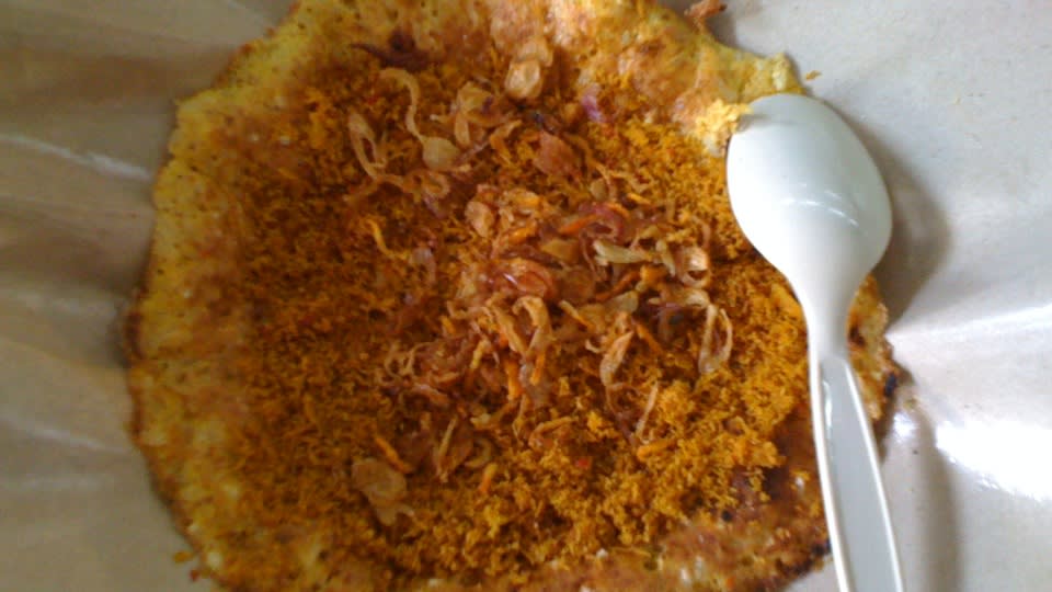 Kerak telor berwarna keemasan dengan taburan serundeng dan bawang goreng di atas kertas pembungkus cokelat dengan sendok plastik putih, siap disantap.