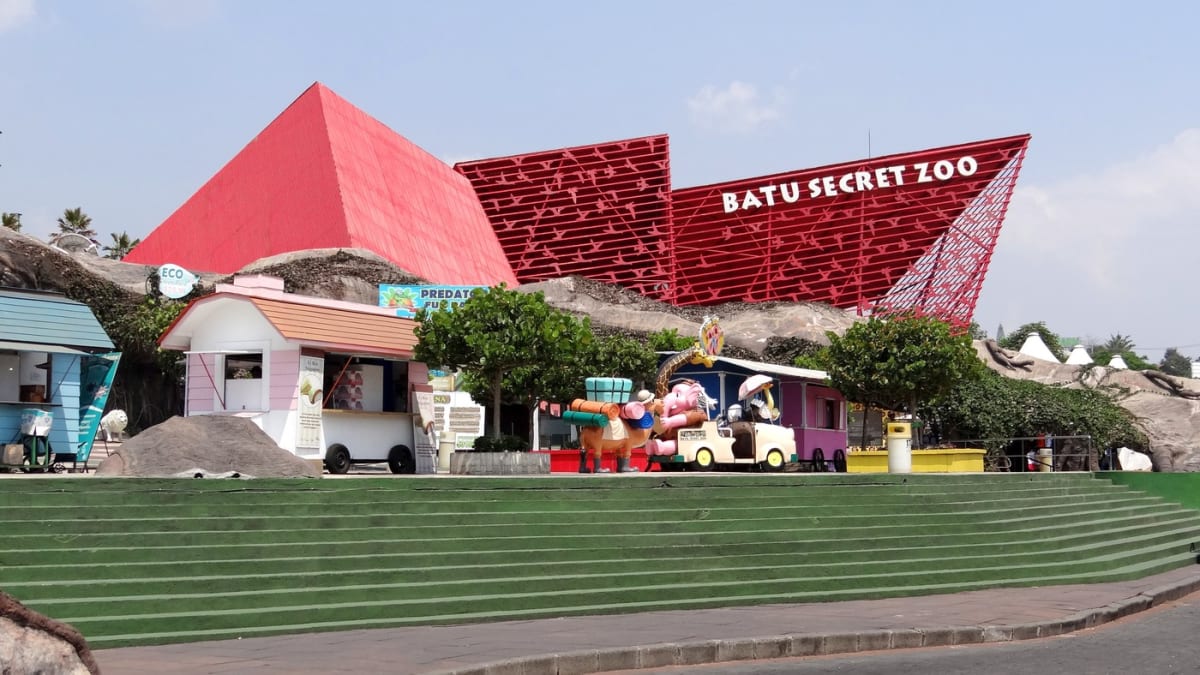 Fasad merah gerbang Batu Secret Zoo bertulisan BATU SECRET ZOO dengan deretan kios berwarna, mobil terbuka berisi figur kartun satwa, unta bermuatan koper di sebelahnya, dan tangga hijau menuju pintu masuk