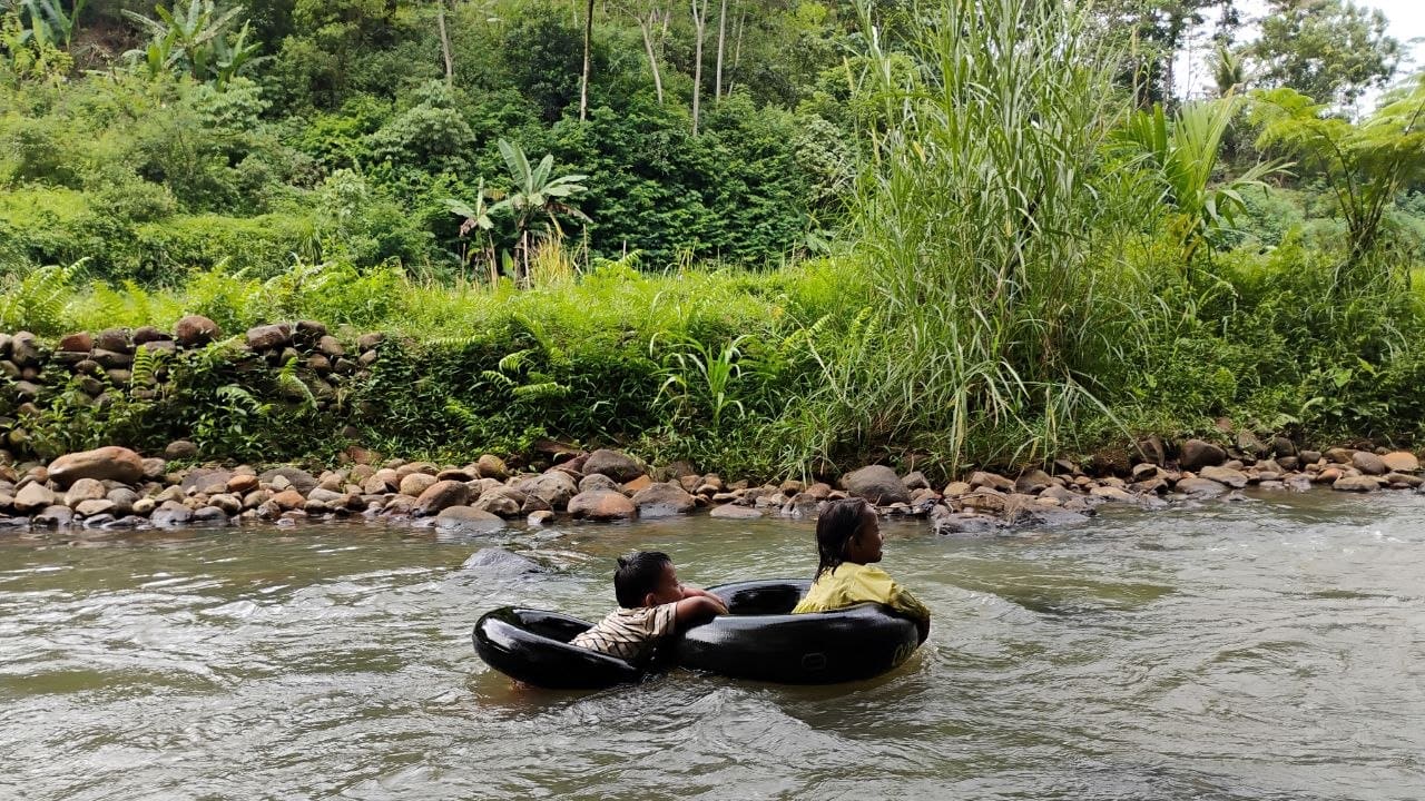 Dua anak bermain menggunakan ban di sungai berbatu yang dikelilingi pepohonan hijau