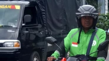 Seorang pengemudi ojek online berhelm hitam dengan jaket kerja hijau duduk di atas sepeda motornya di jalan, dengan mobil box gelap di belakangnya.