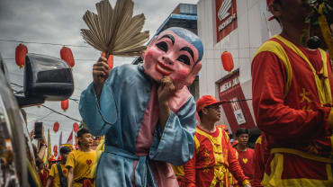 Peserta pawai Cap Go Meh di Singkawang berjalan di jalan kota pada siang hari, mengenakan kostum tradisional warna merah dan kuning serta topeng besar merah muda dan biru, dengan lampion merah menggantung di atas jalan